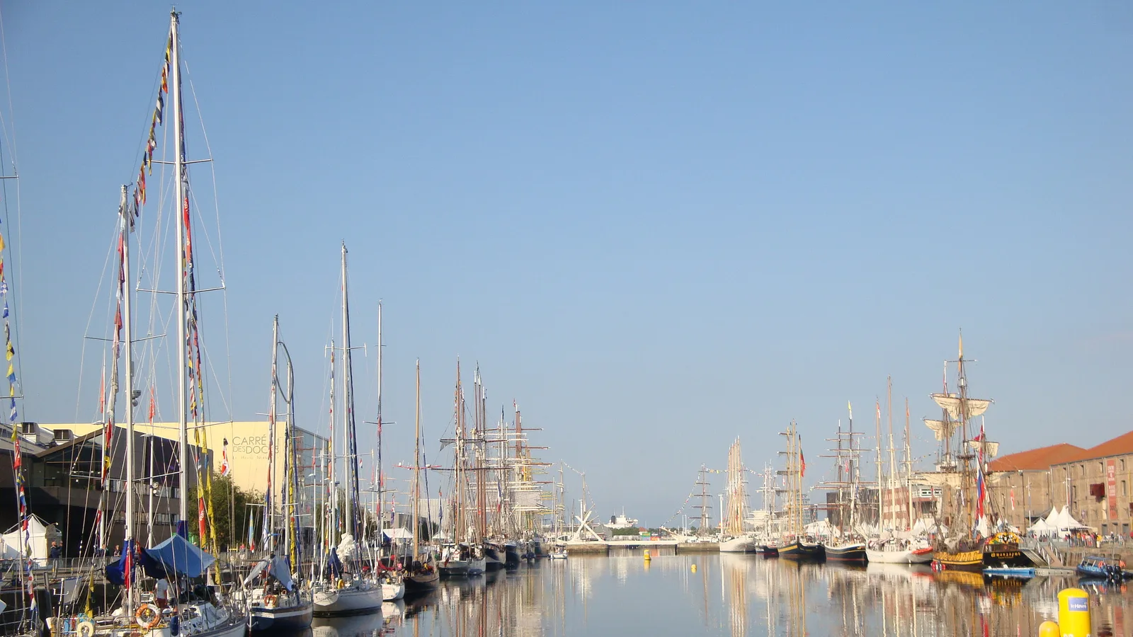 Tall ships and yachts moored in a Le Havre basin and reflected in still water on a clear morning.