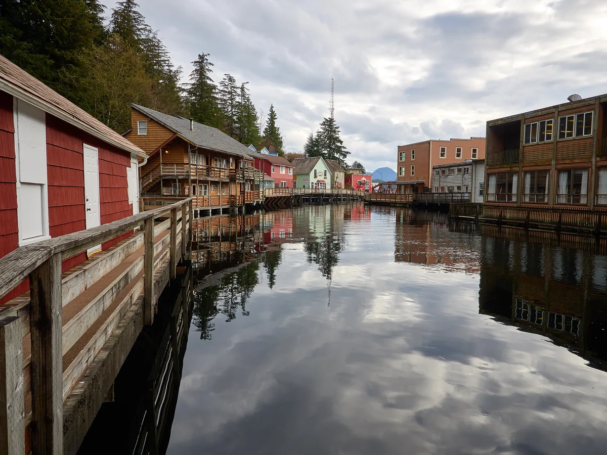 Still water of Ketchikan Creek reflecting clouds, with red wooden buildings and the Creek Street boardwalk along the bank.