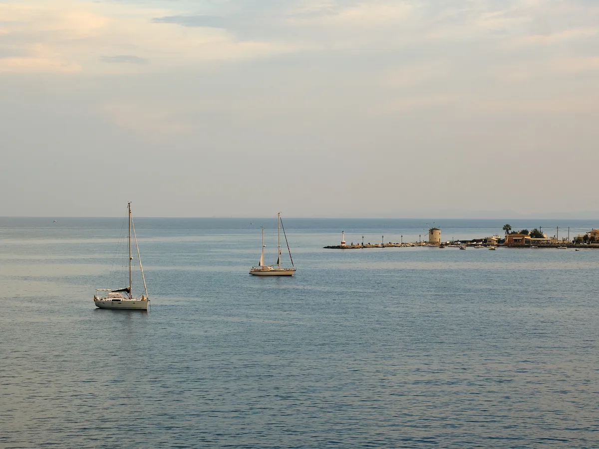 Two sailboats on calm hazy water of the Bay of Garitsa with a jetty and windmill-topped point in the distance.