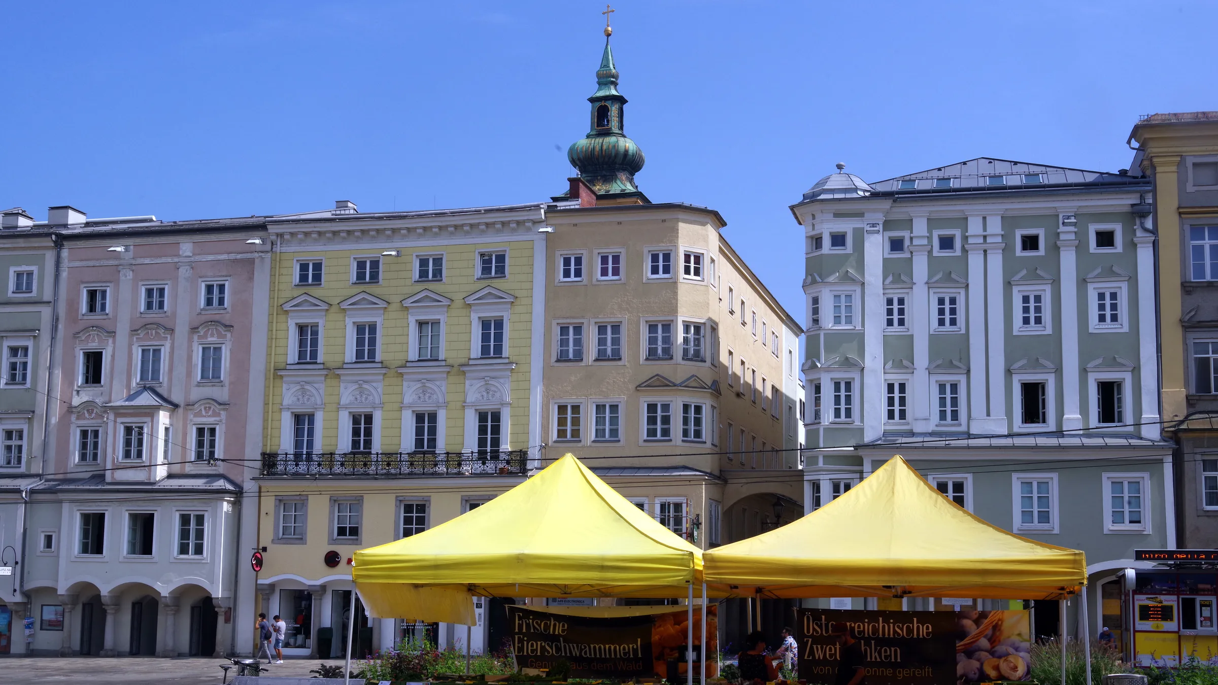 The Linz Hauptplatz on a market day with yellow stalls in front of pastel baroque houses.