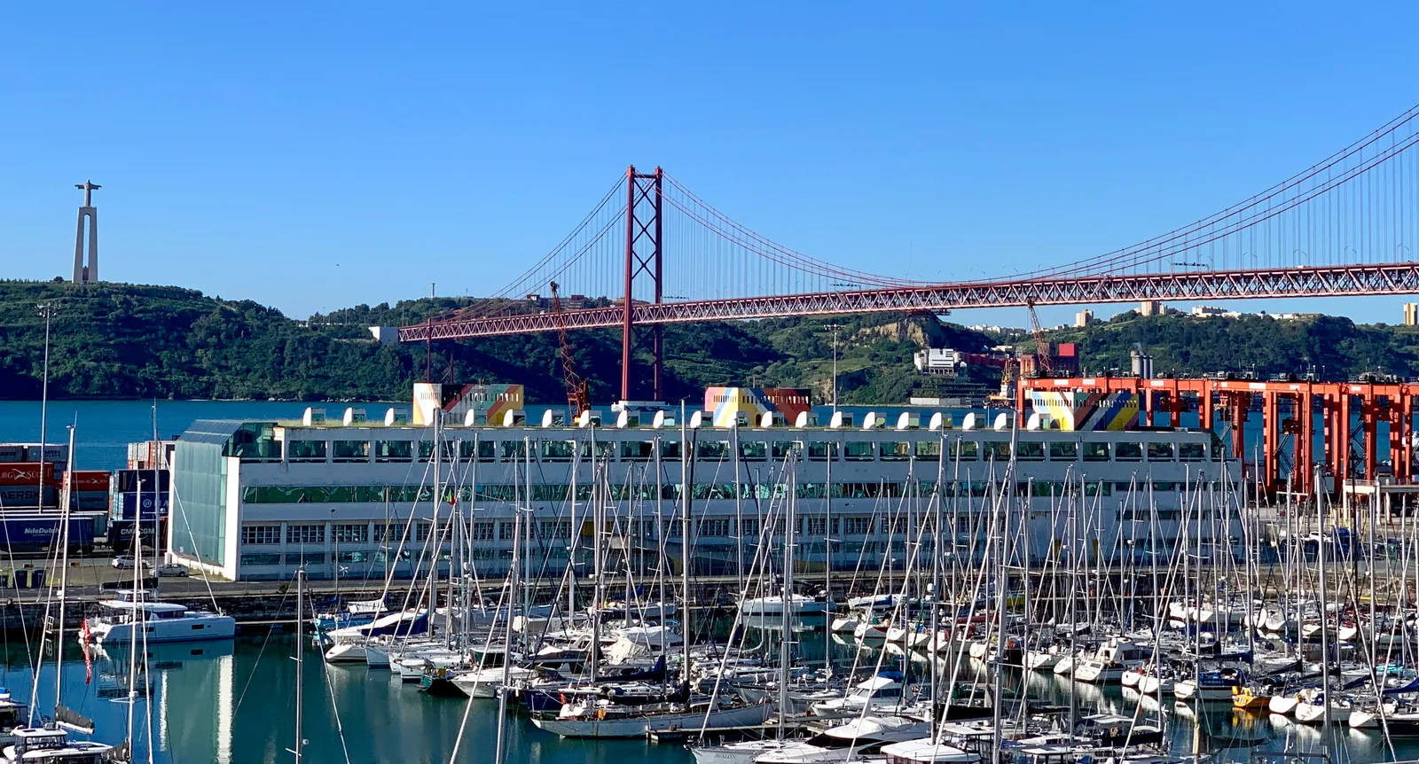 Yachts in a marina below the 25 de Abril Bridge with the Cristo Rei hill beyond.