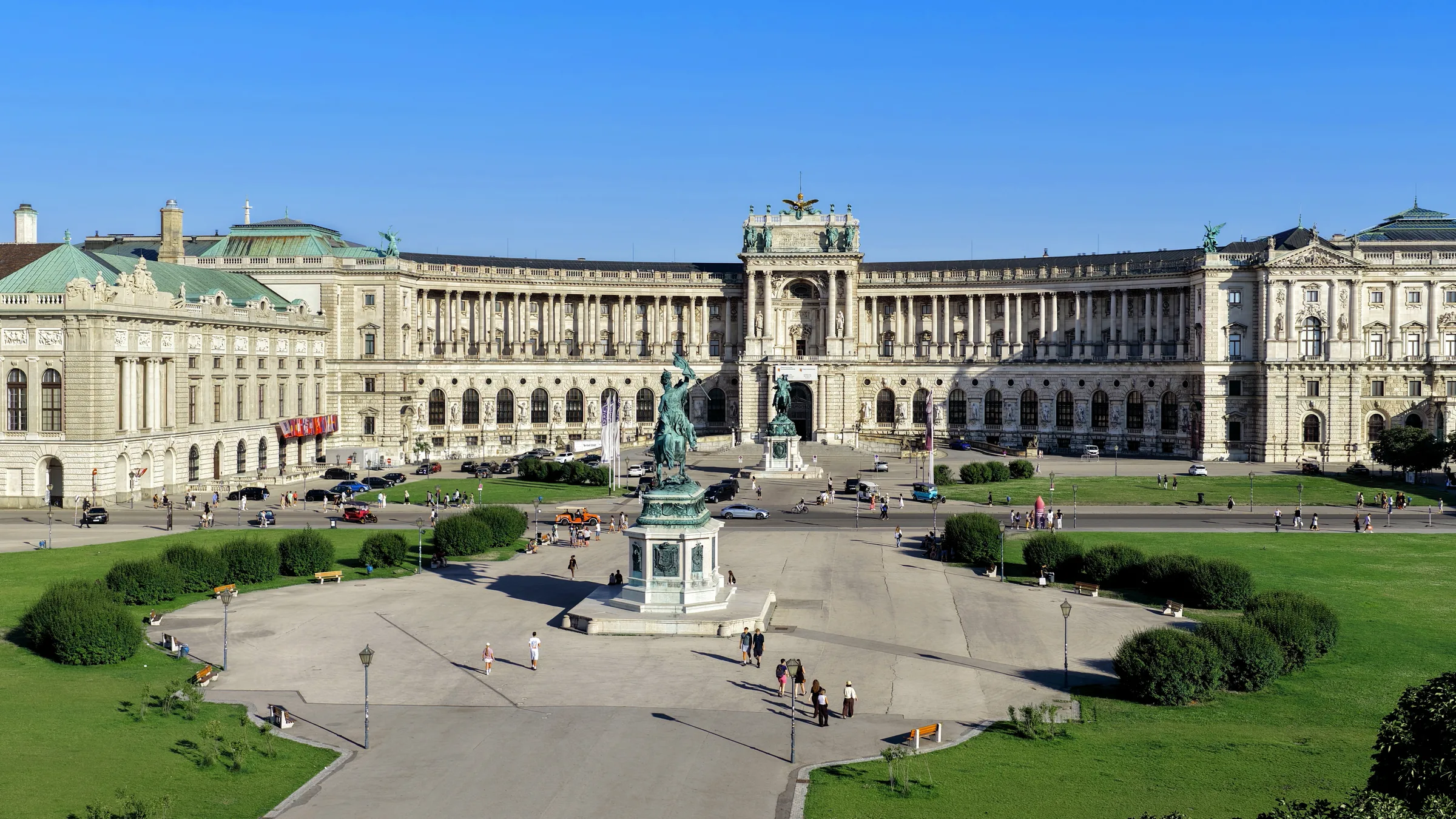 A curved palace front behind the open lawns and paths of the Heldenplatz in Vienna, under a clear sky.