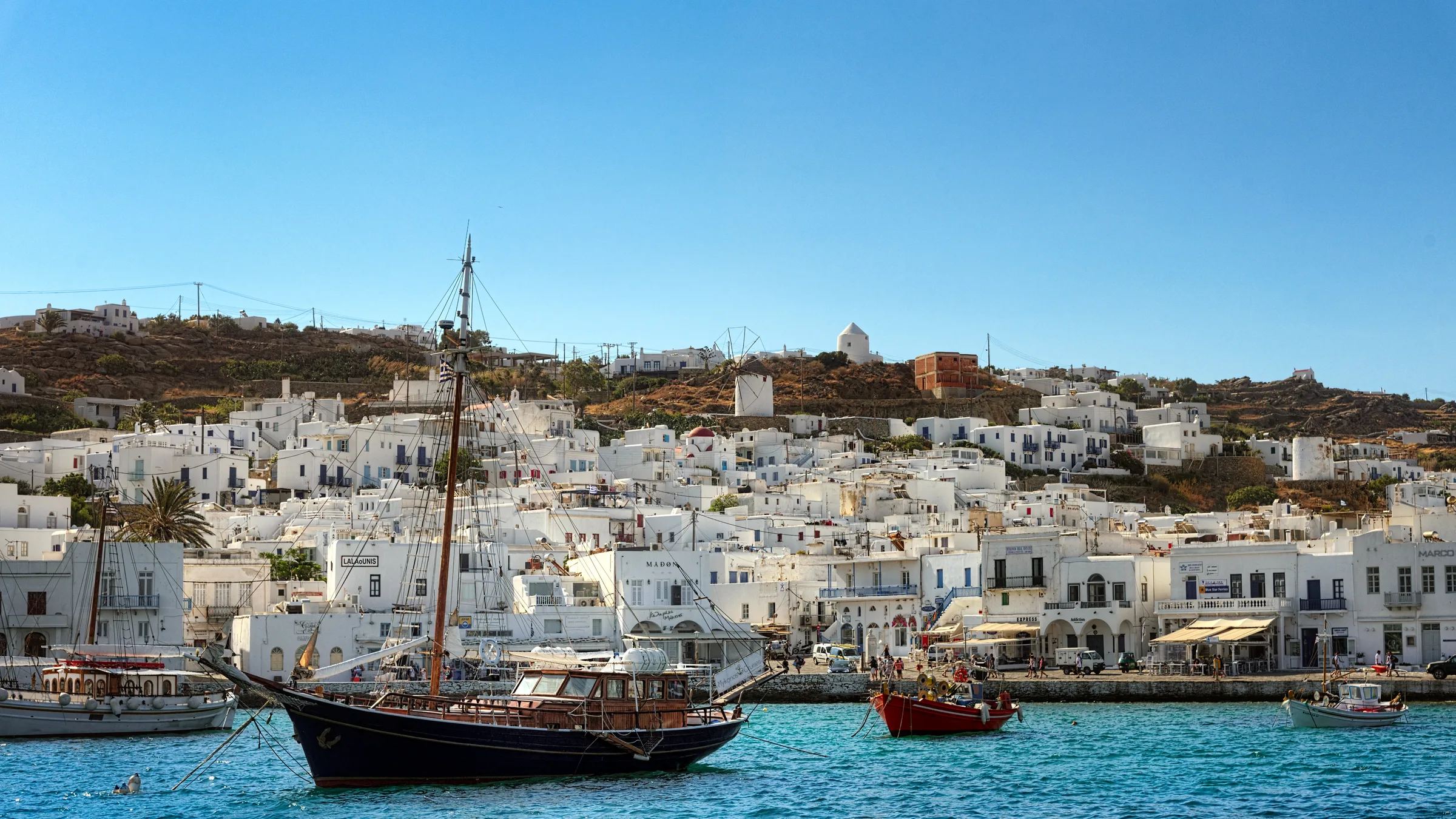 A dark-hulled sailing boat moored in the old harbour of Mykonos Town with whitewashed buildings and a windmill on the ridge.