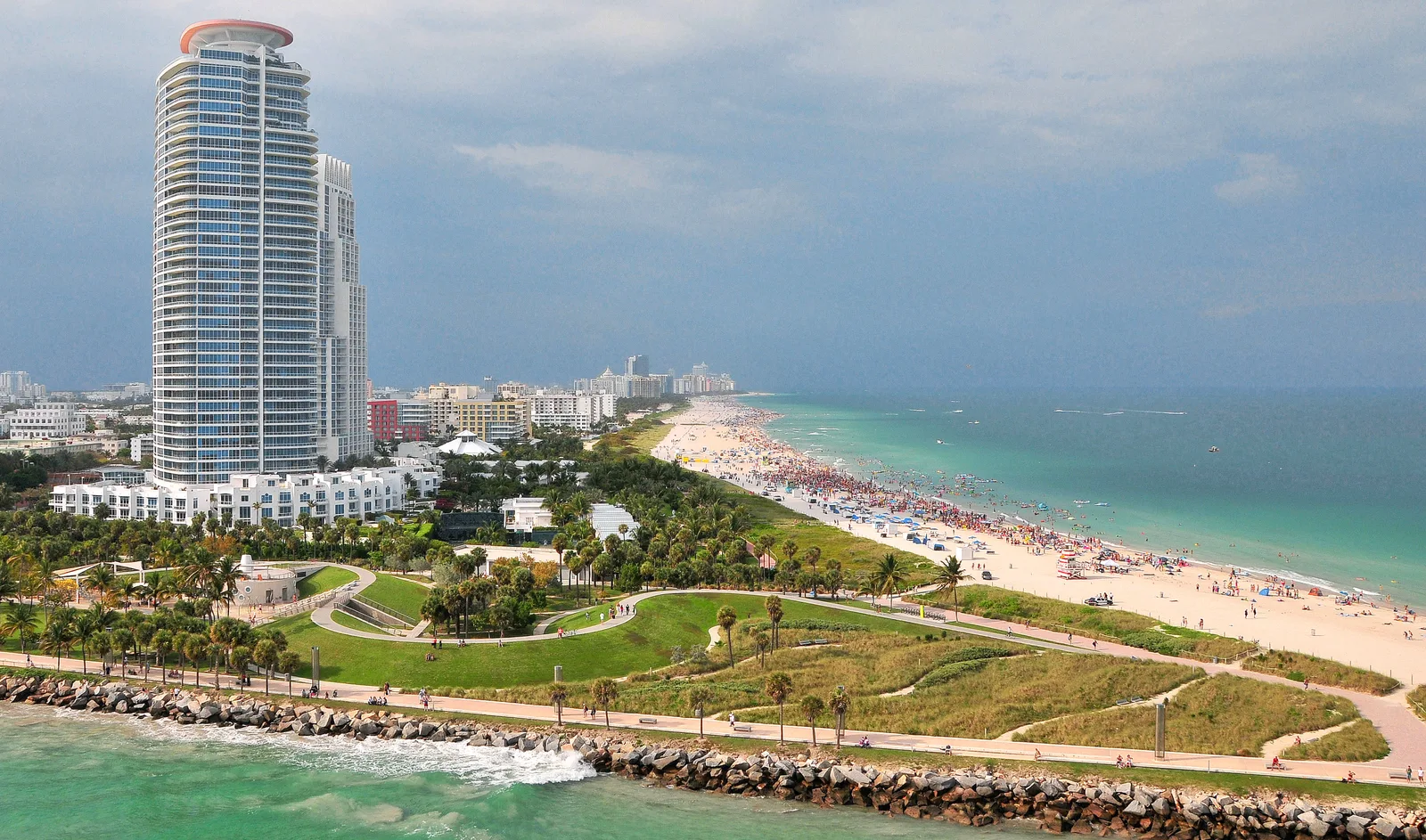 Aerial view of South Pointe Park, a rock jetty and a wide beach with a tall residential tower and an approaching storm.