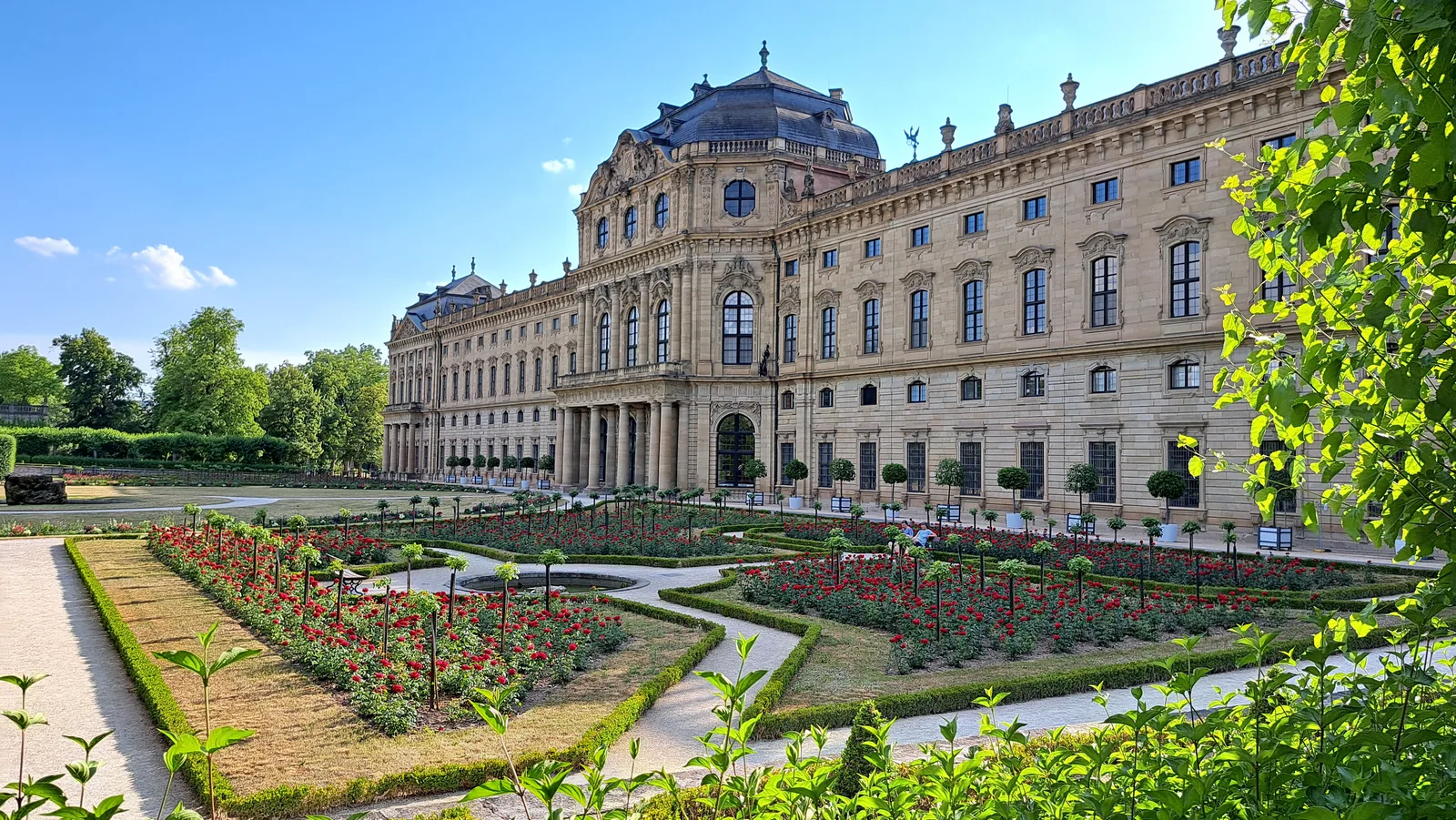 The Würzburg Residence palace and its formal garden with flower beds, in bright sun.