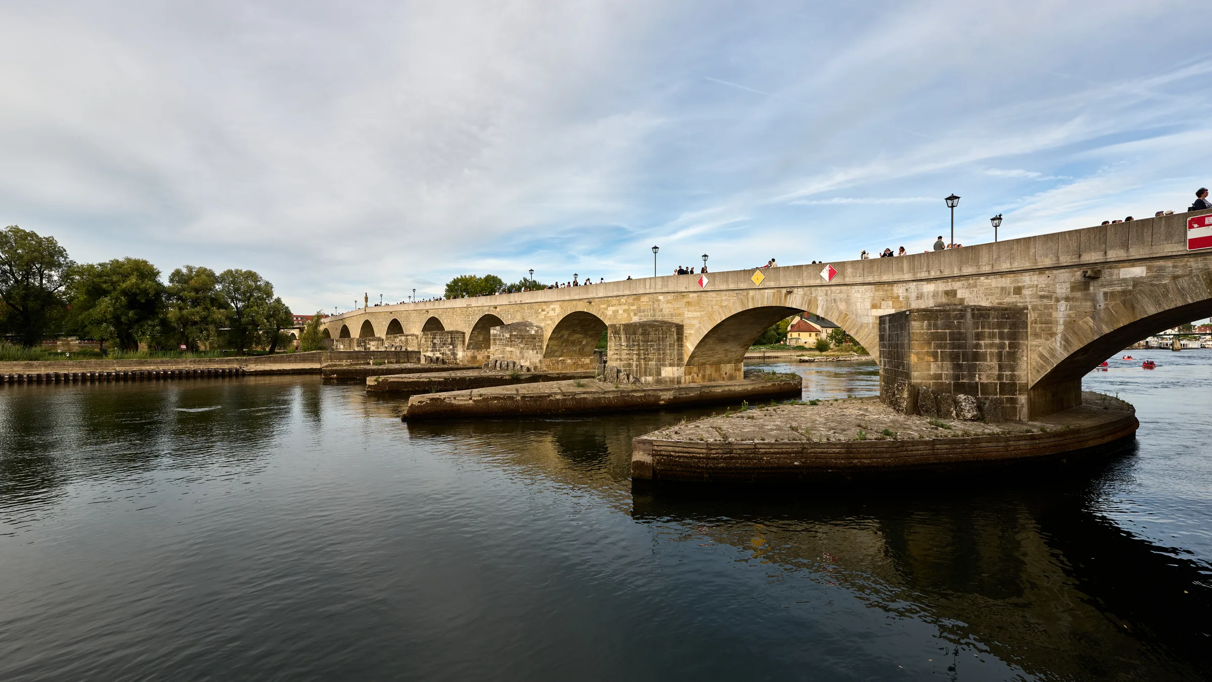The Steinerne Brücke stone bridge over the Danube in Regensburg under a high sky with still water in the foreground.