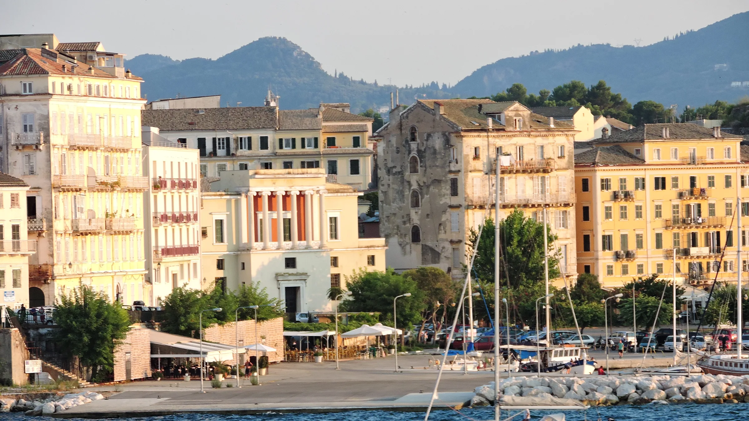 Venetian-style pastel buildings along the old port of Corfu Town with a sailing boat on the water and wooded hills behind.
