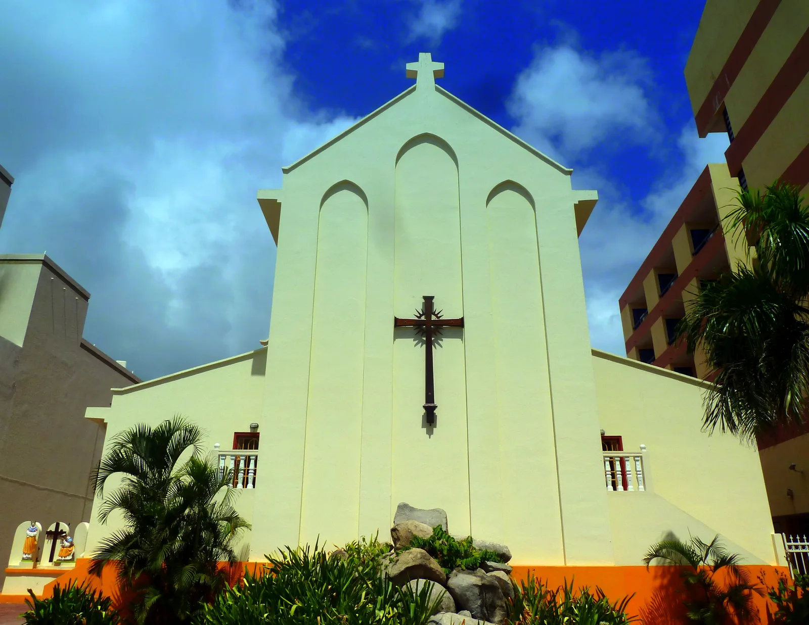 A pale yellow Catholic church with a tall cross on its facade stands among palms and apartment blocks in Philipsburg.