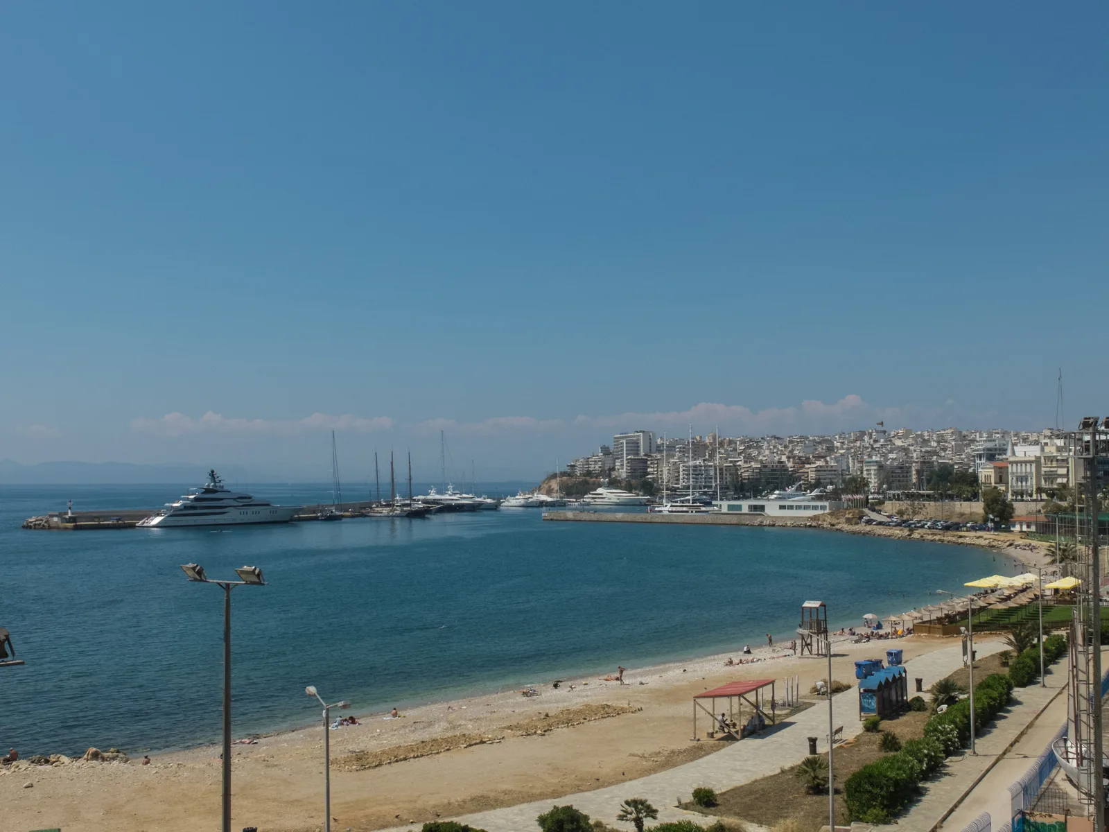 A curved sandy shore beside Piraeus with a megayacht at a breakwater and the city climbing the hillside.