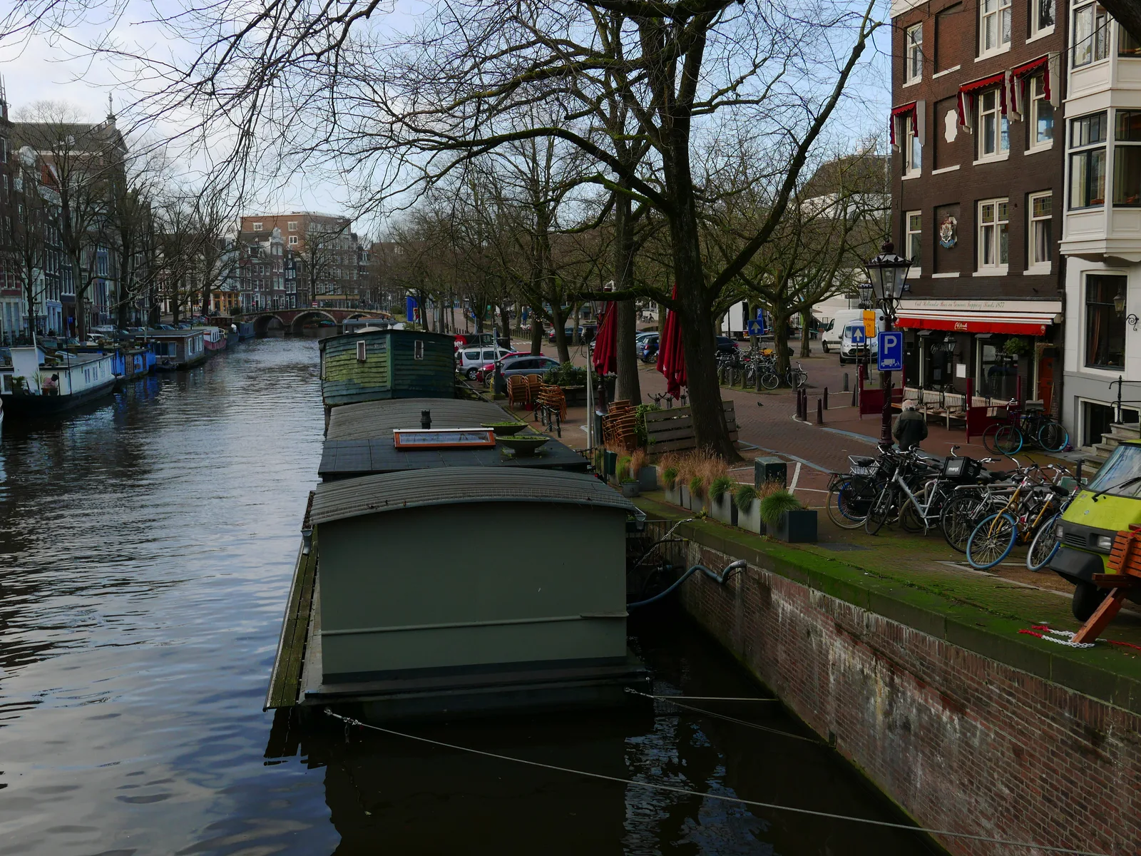 A calm Amsterdam canal with a moored houseboat, brick quay, bicycles and bare trees along Prinsengracht.