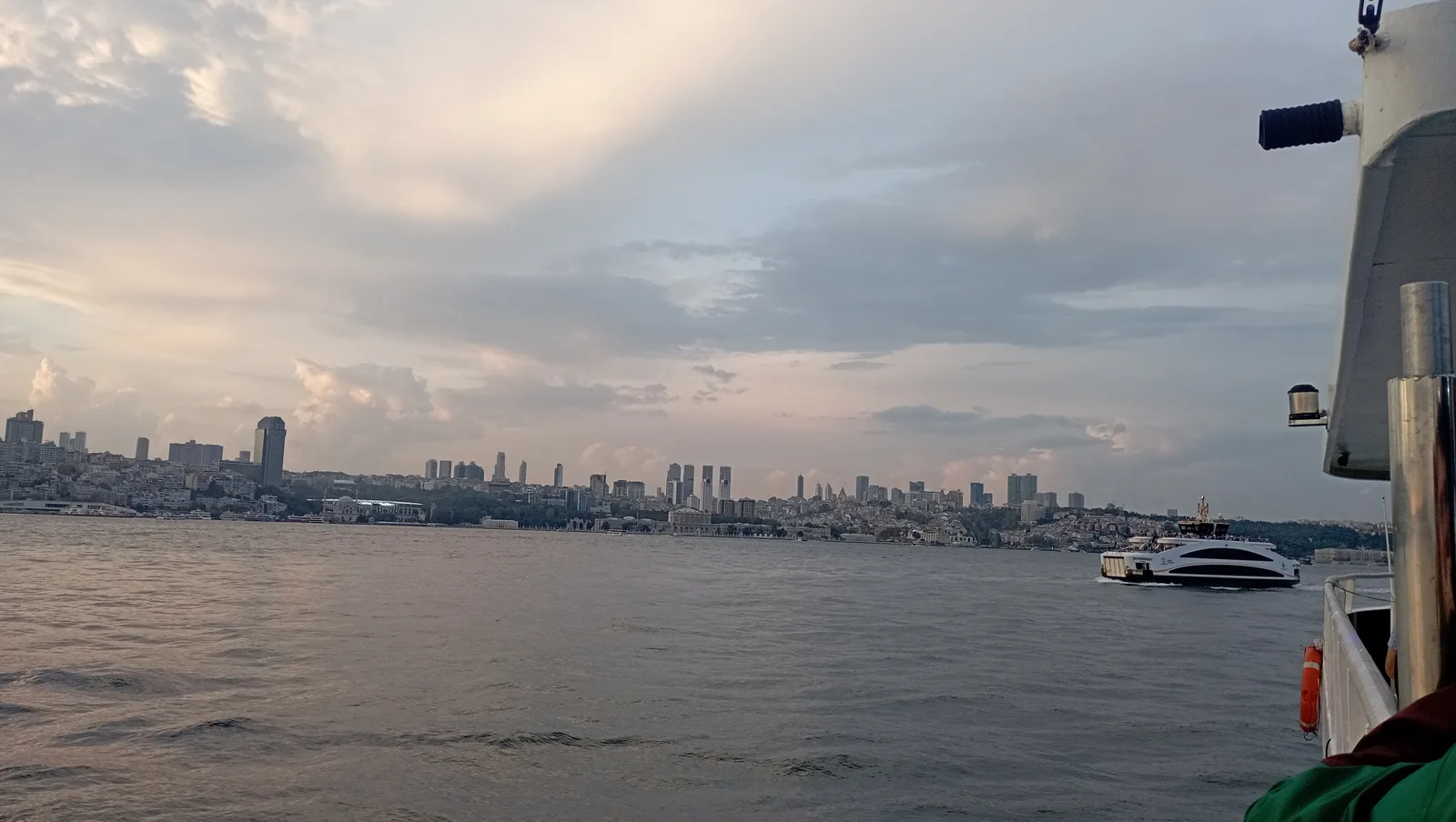 A grey-pink evening sky over the Bosphorus with the city skyline in the distance and a ferry deck edge at right.