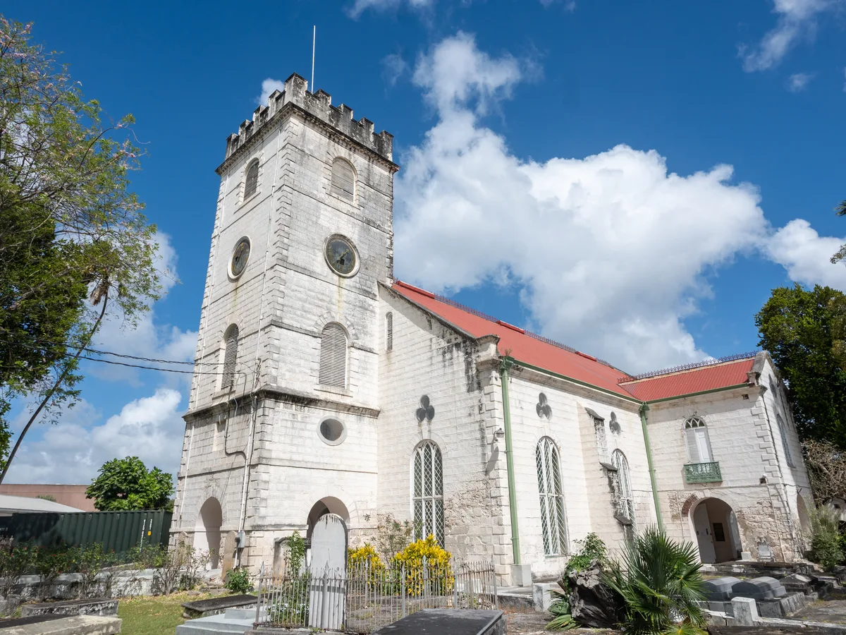 A white stone church with a square clock tower and red roof stands against a blue sky in Bridgetown.