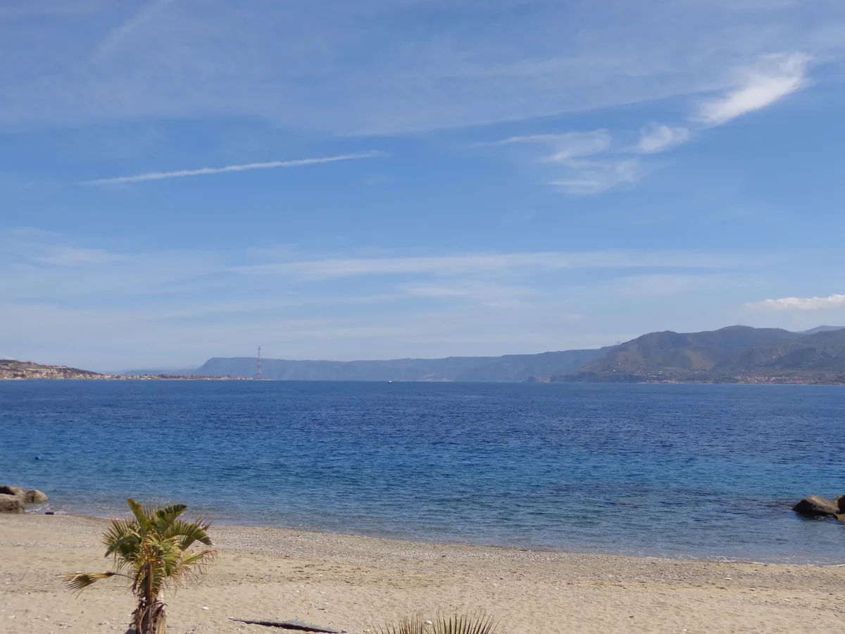 A pale sand beach on the Strait of Messina with a palm in the foreground and the opposite coast across the water.
