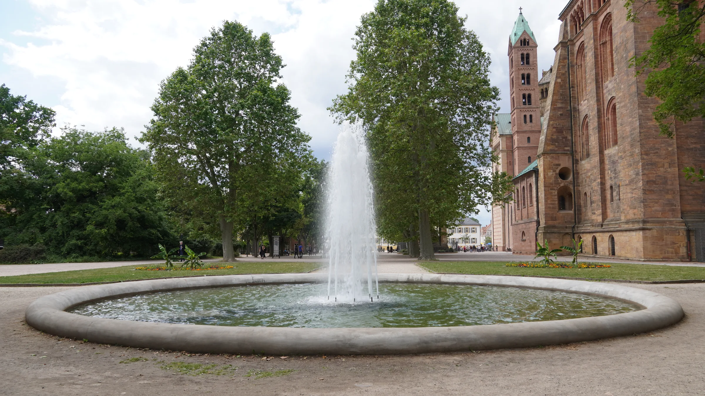 A round fountain in the garden by Speyer cathedral, with the red sandstone cathedral behind the trees.