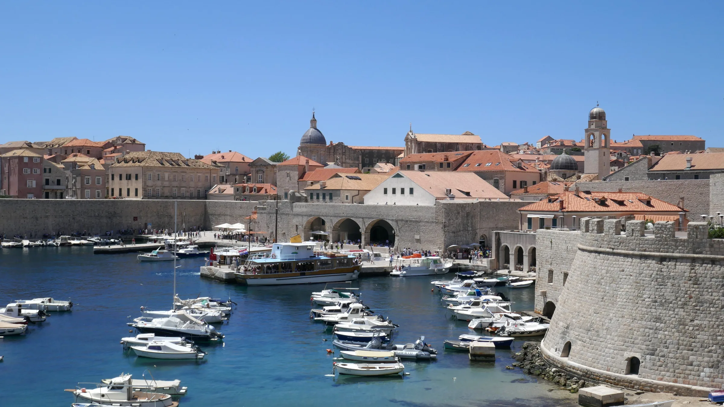 The old harbour of Dubrovnik with moored boats under the city walls, a bell tower and a cathedral dome above terracotta roofs.