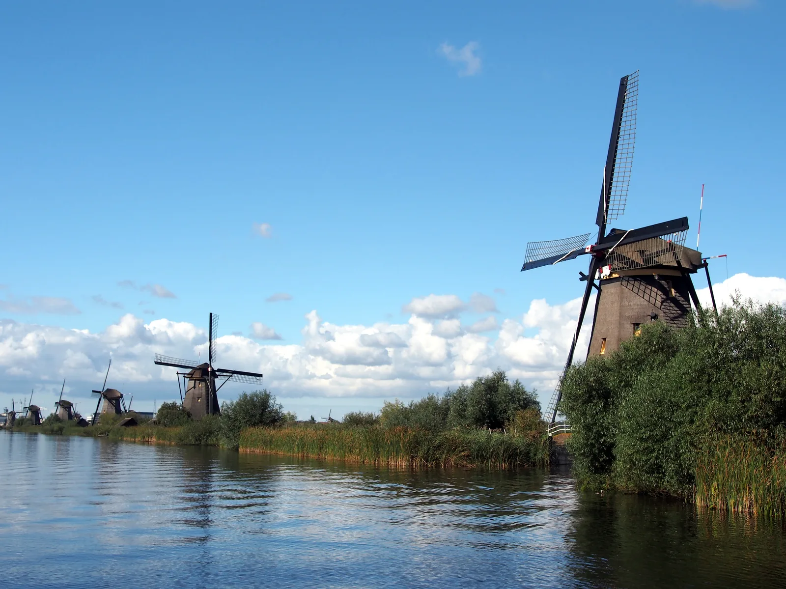 A windmill by the canal at Kinderdijk with a clear blue sky and white clouds above a stretch of still water.
