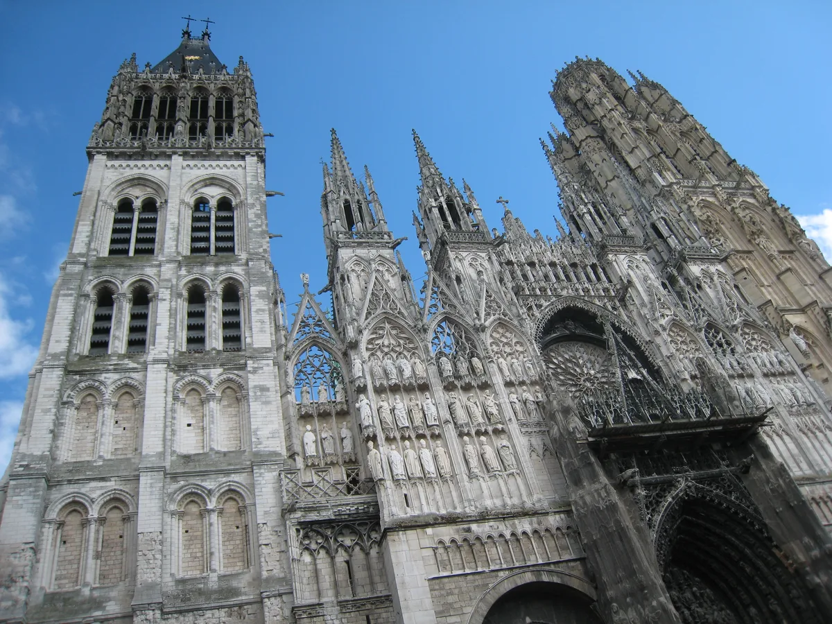 The pale carved towers and facade of the cathedral in Rouen seen from below against a blue sky.
