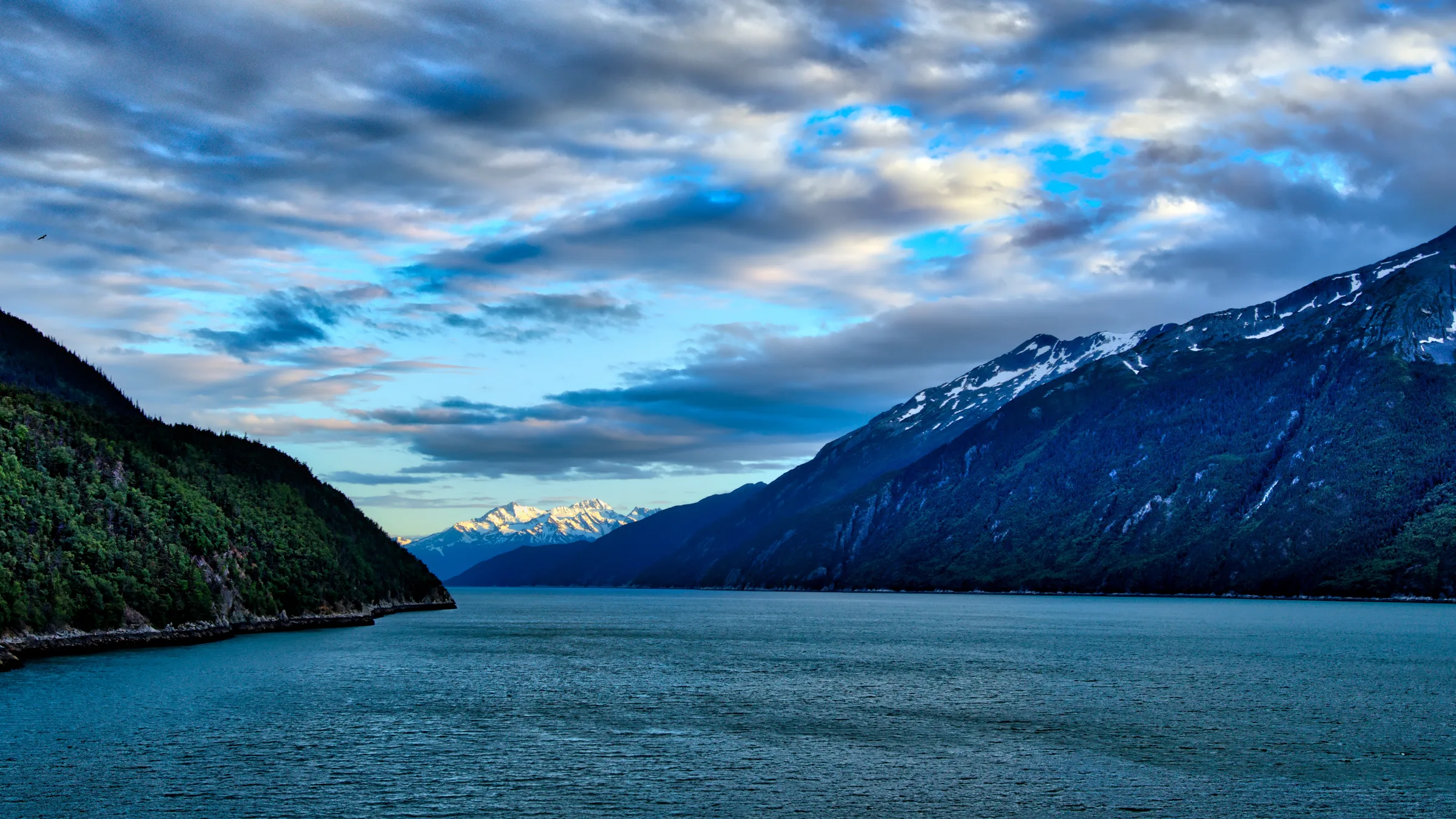 A long fjord arm near Skagway with dark mountains, snow patches and a dramatic cloud-streaked sky.