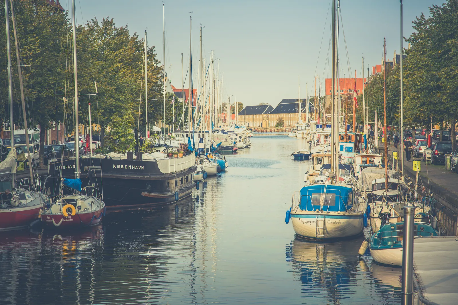 A calm canal in Christianshavn in Copenhagen lined with moored sailboats and a black houseboat.