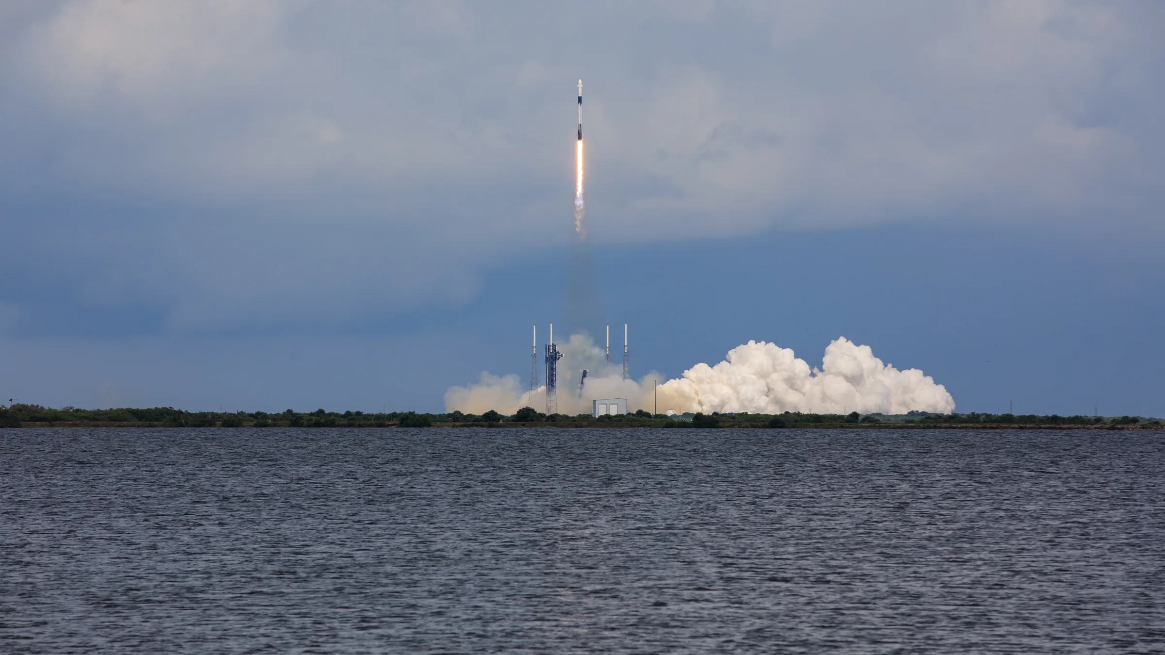 A rocket lifts off from a distant launch pad, with a white cloud of exhaust below it and dark water in the foreground.