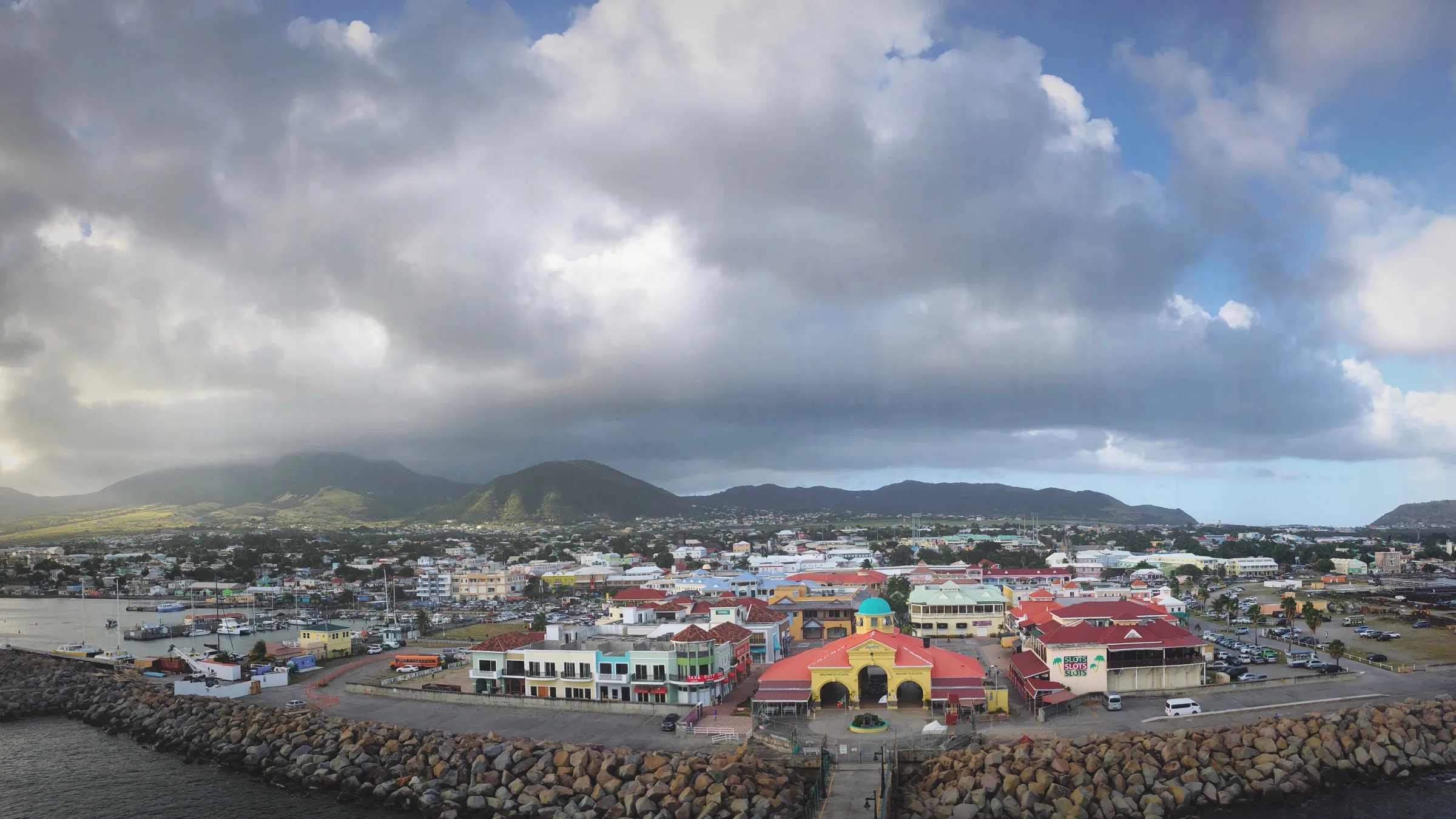 A sweeping view of Basseterre and the cruise terminal area with the town's roofs below the green mountains of St Kitts under a big cloudy sky.