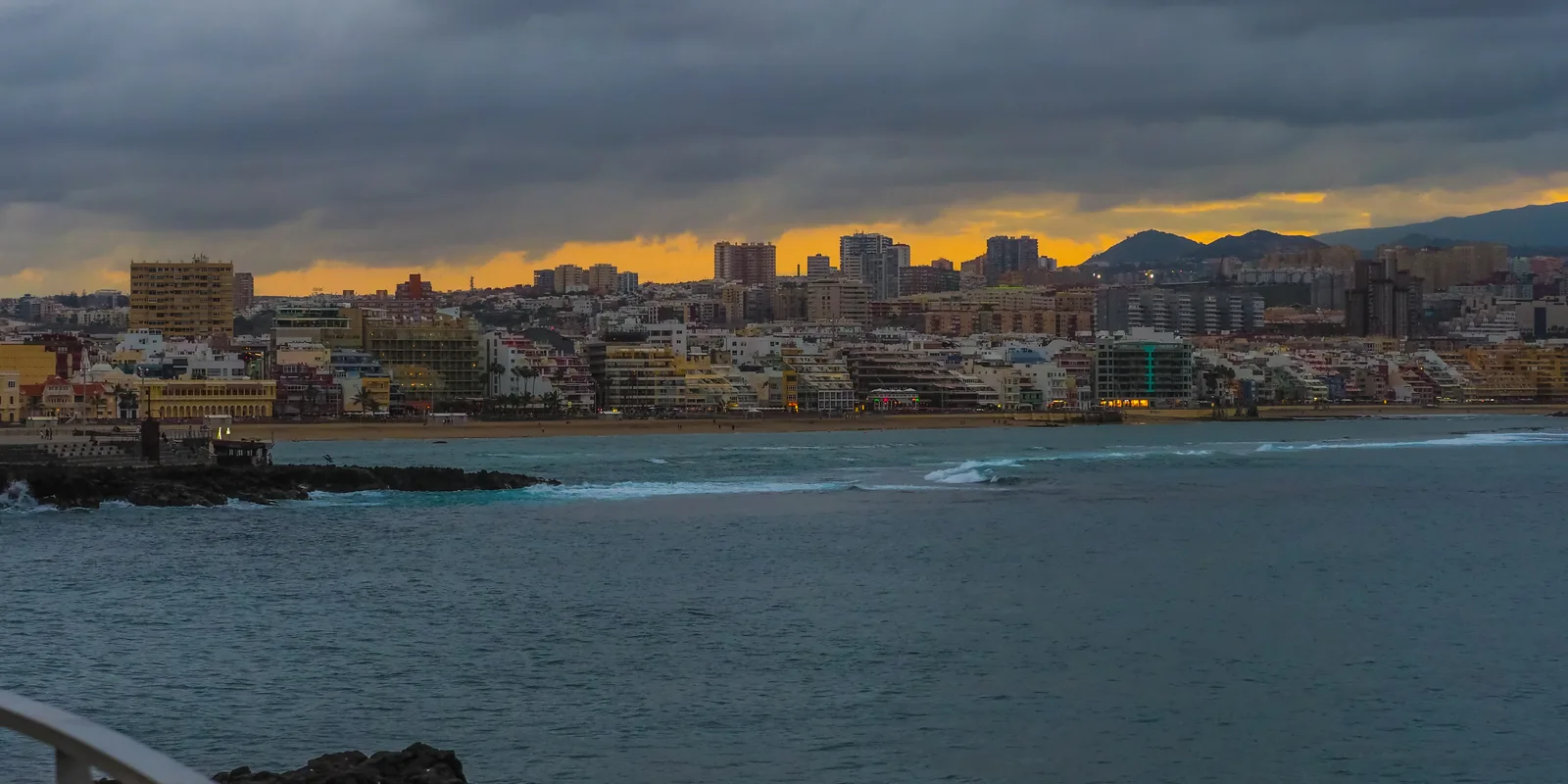 The Las Palmas seafront and Las Canteras bay under a dark stormy sky with a band of orange sunset light on the horizon.