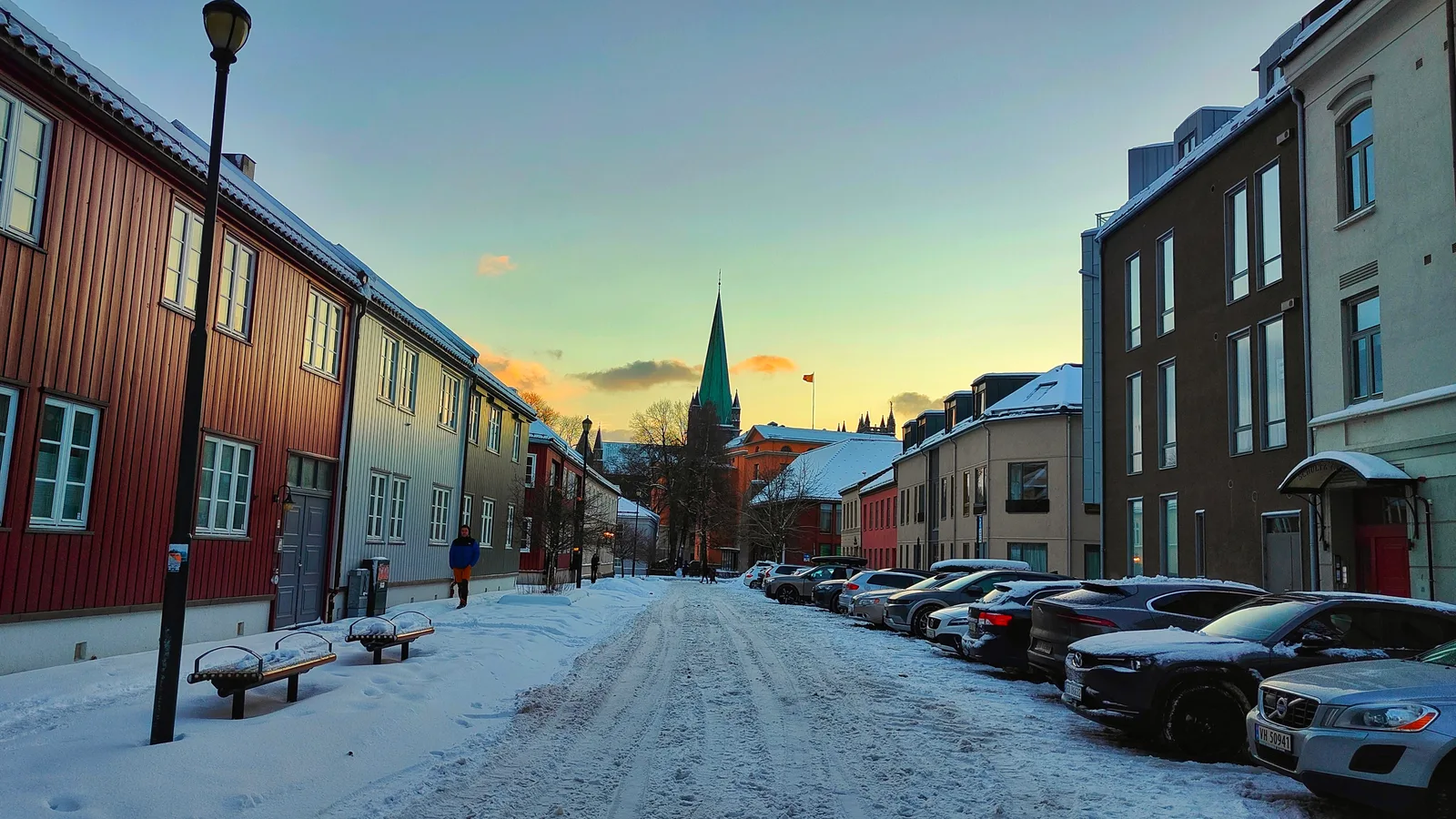 A snowy Trondheim street at dusk with timber warehouses and the Nidaros Cathedral spire in the distance.