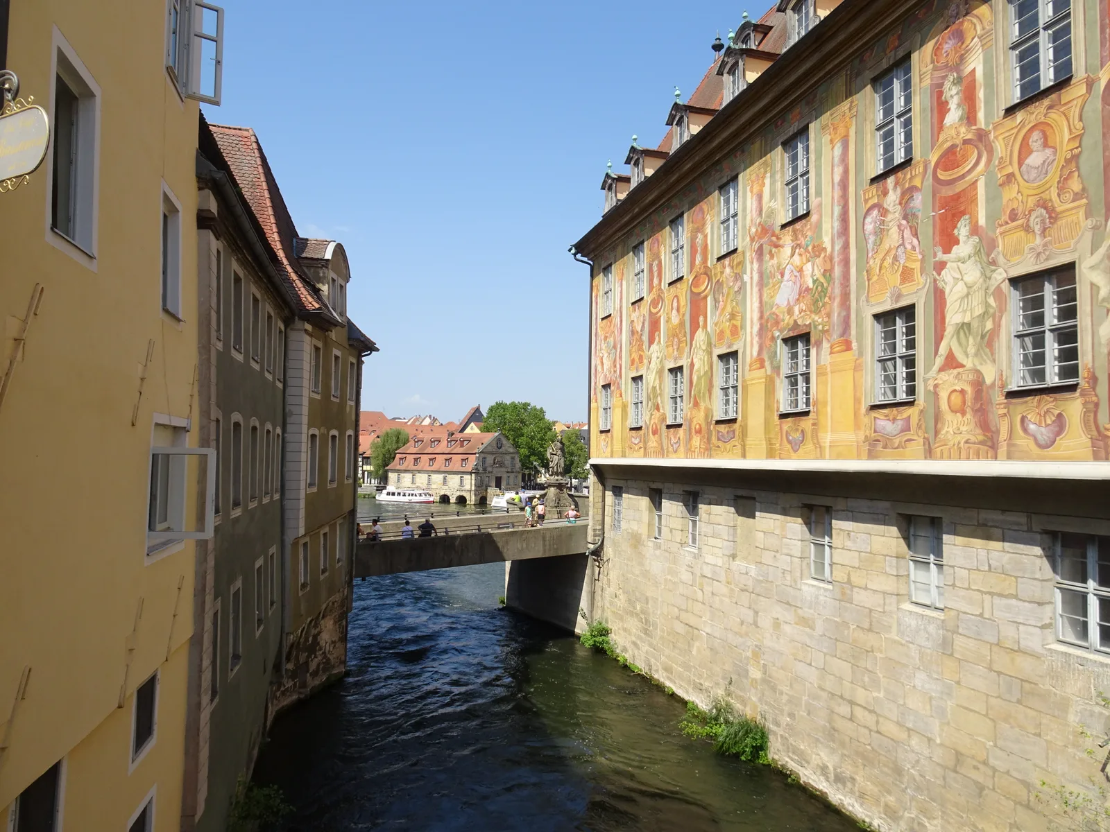 A canal between old houses in Bamberg with a painted facade at the right and a footbridge in the distance.