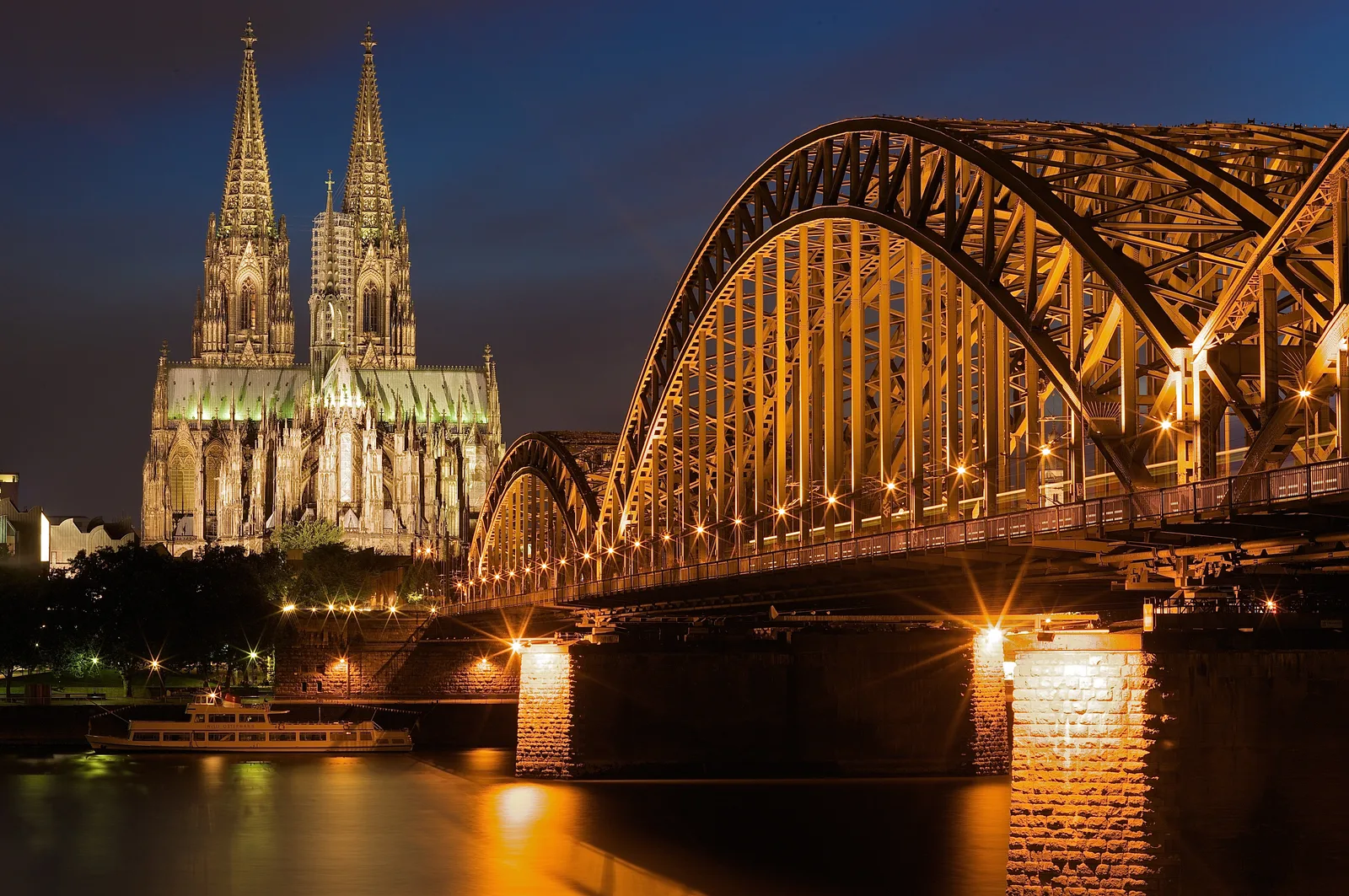 The twin spires of Cologne Cathedral lit at night beside the steel arches of the illuminated Hohenzollern Bridge.
