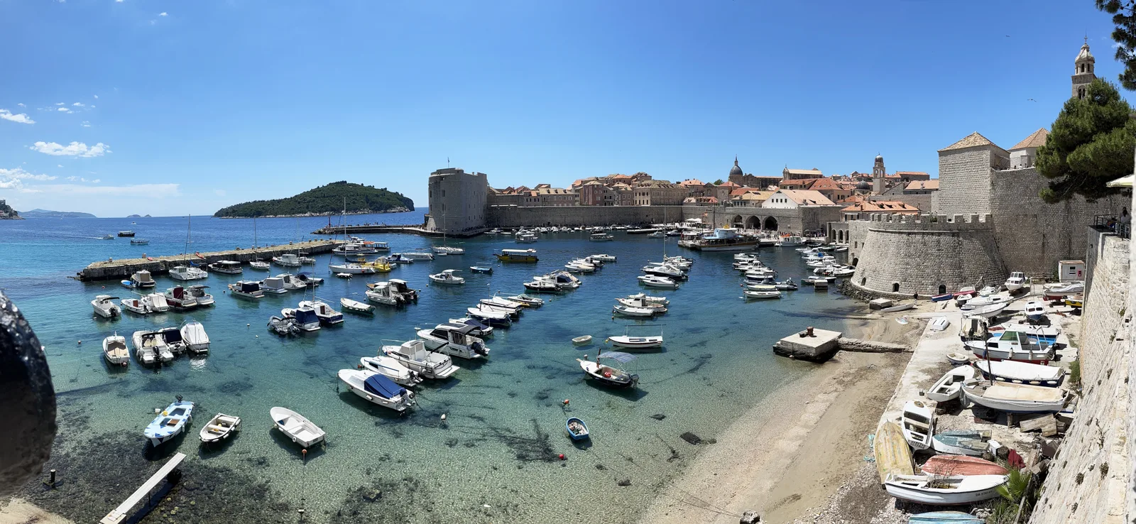 A wide view over the clear water of Dubrovnik's old harbour with many small boats, the walls and the island of Lokrum behind.