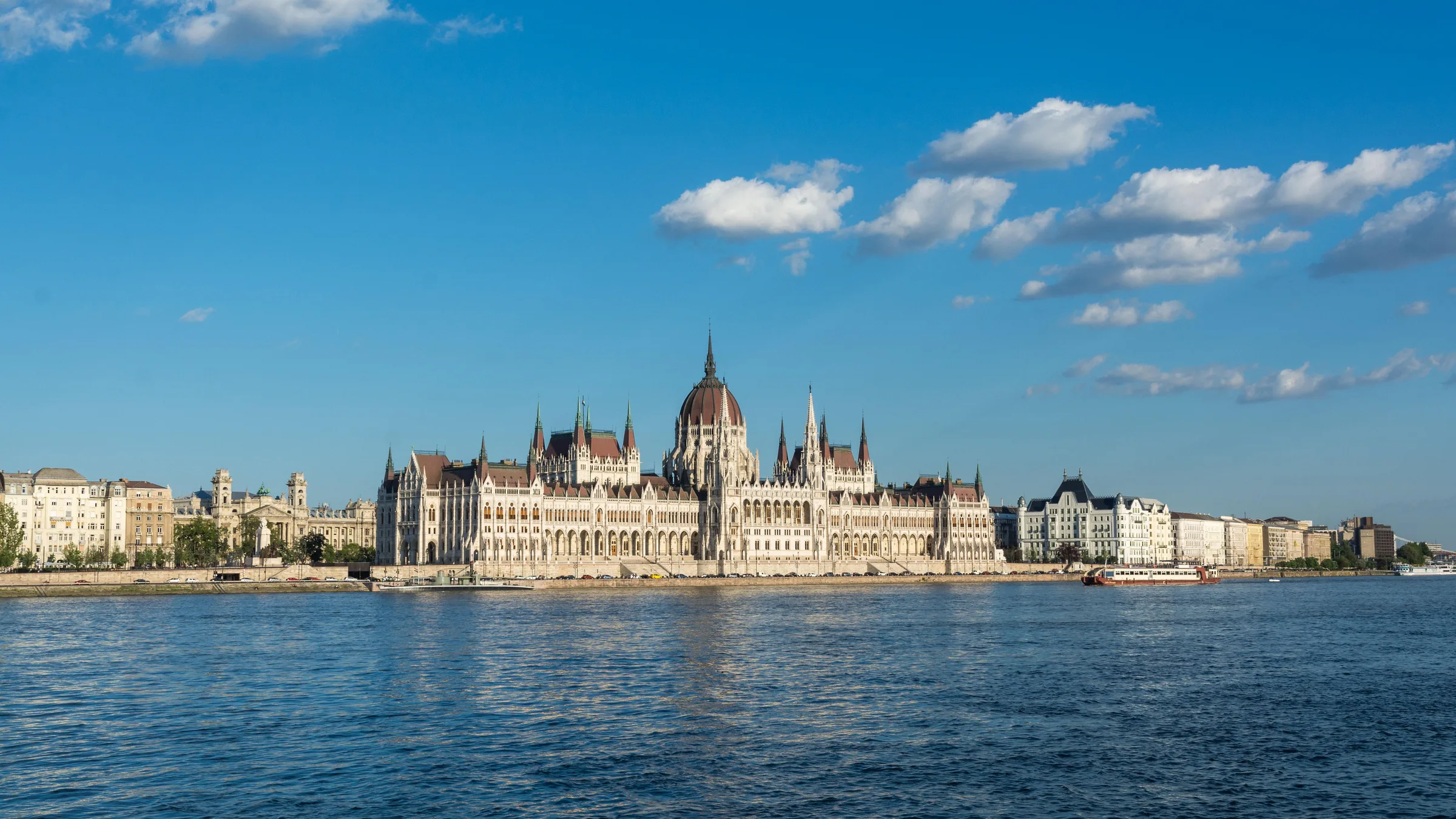 The Hungarian Parliament Building across the Danube on a clear day, with the river in the foreground and Pest buildings on both sides.