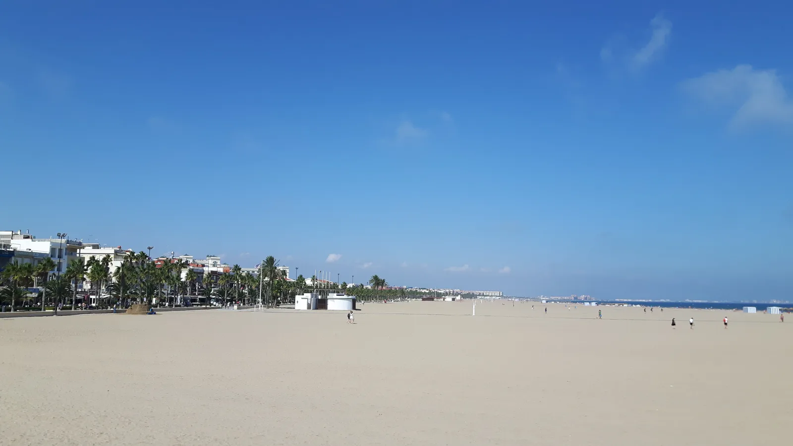 An empty sweep of sand on Malvarrosa beach in Valencia with the seafront promenade under a clear blue sky.