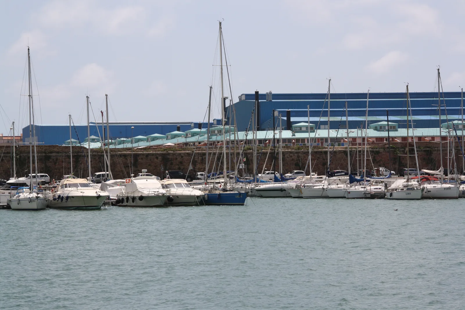 Rows of moored boats in a Livorno marina below a large blue warehouse and old harbour wall.
