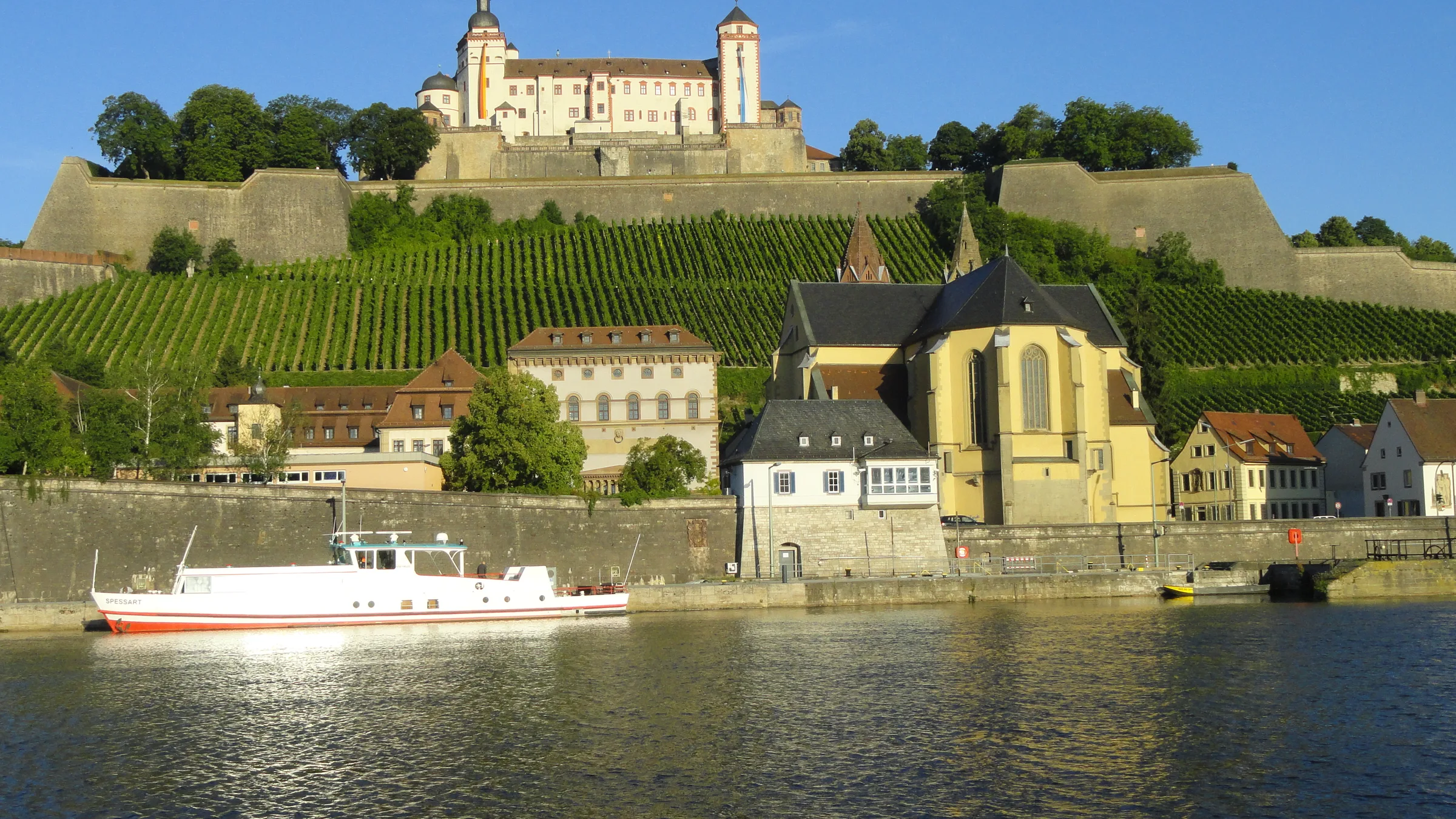 The Marienberg Fortress above vineyard terraces and a river boat on the Main in Würzburg in bright light.