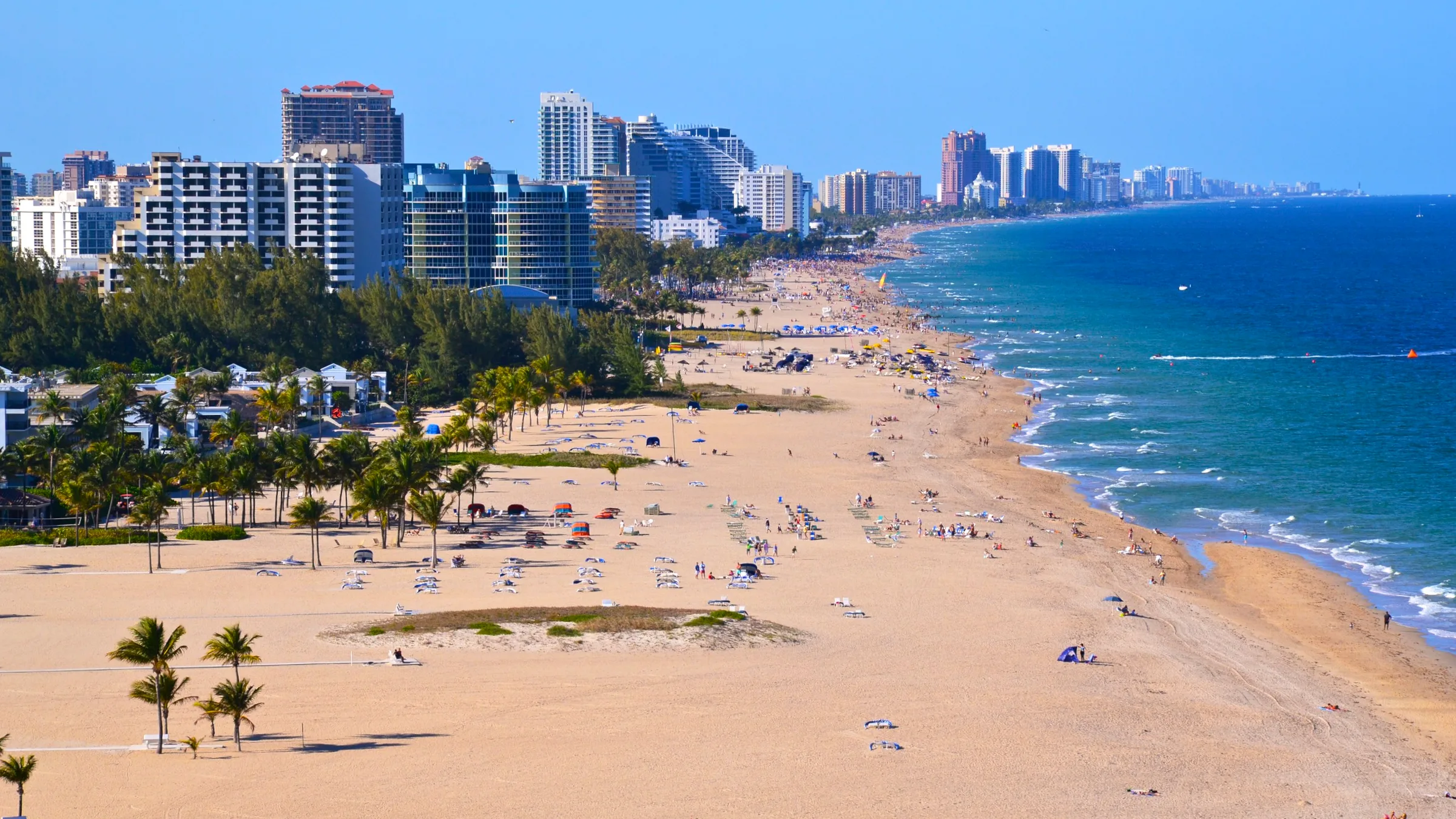 Aerial view along Fort Lauderdale beach with a long curve of sand, blue surf and a row of high-rise buildings.