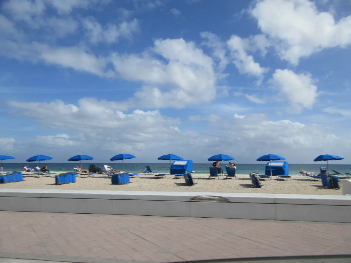 Rows of blue beach umbrellas on the sand under a sky of scattered clouds, seen from the A1A beachfront promenade.