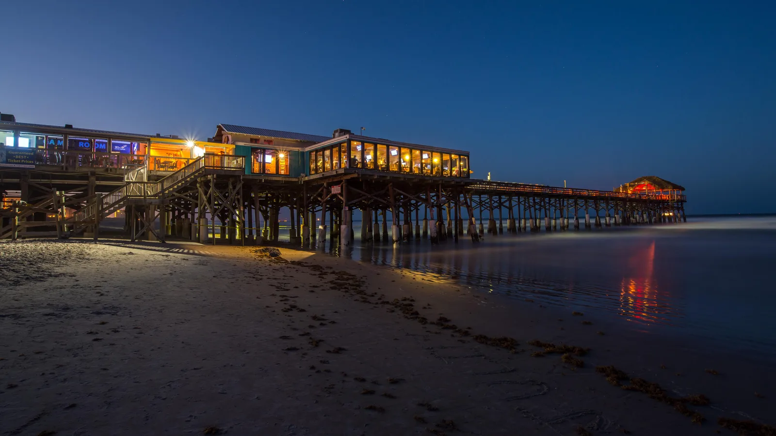 Cocoa Beach Pier lit from within at blue hour, with the pier reflected on wet sand.