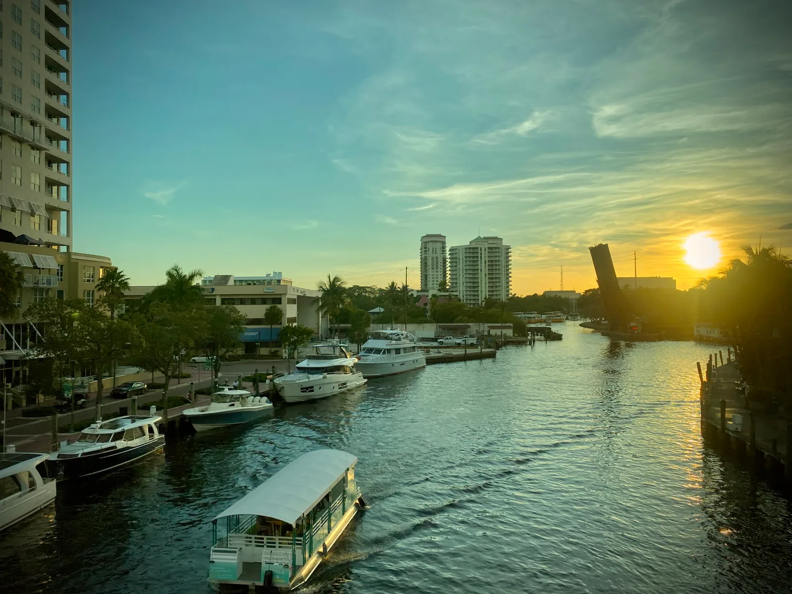 Boats moored along a Fort Lauderdale waterway as the sun sets behind a raised drawbridge, with a small water taxi in the foreground.