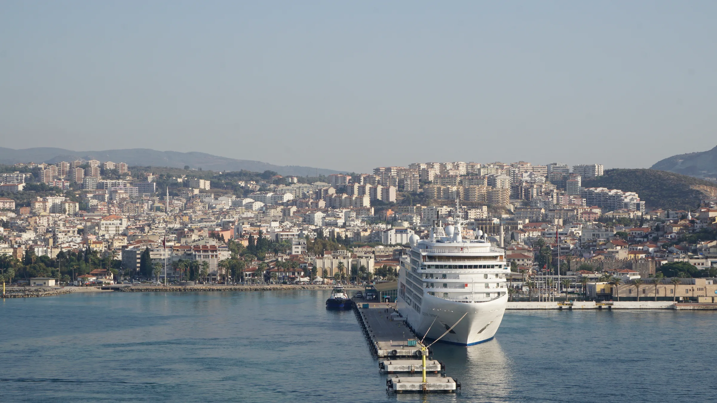 A white cruise ship berthed at a floating pier in Kusadasi harbour with the dense town and hills behind under a hazy sky.