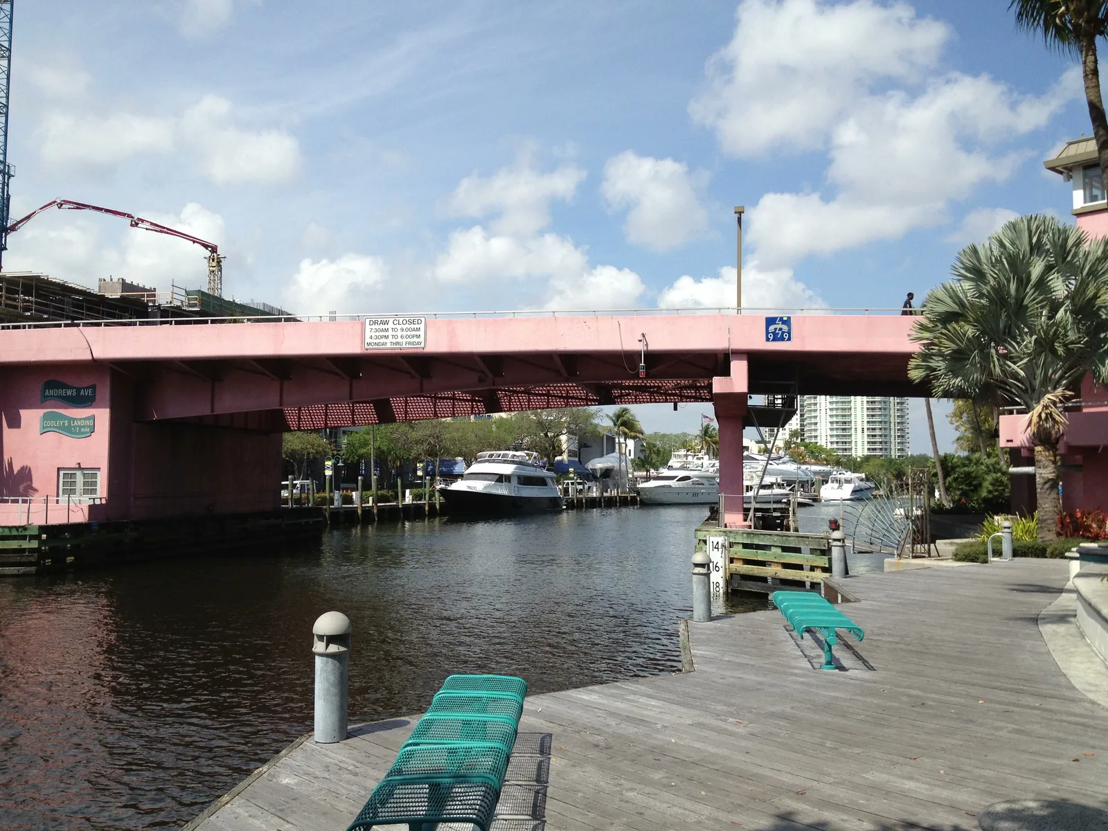 A pink drawbridge span over the New River from the Riverwalk boardwalk, with boats and green benches.