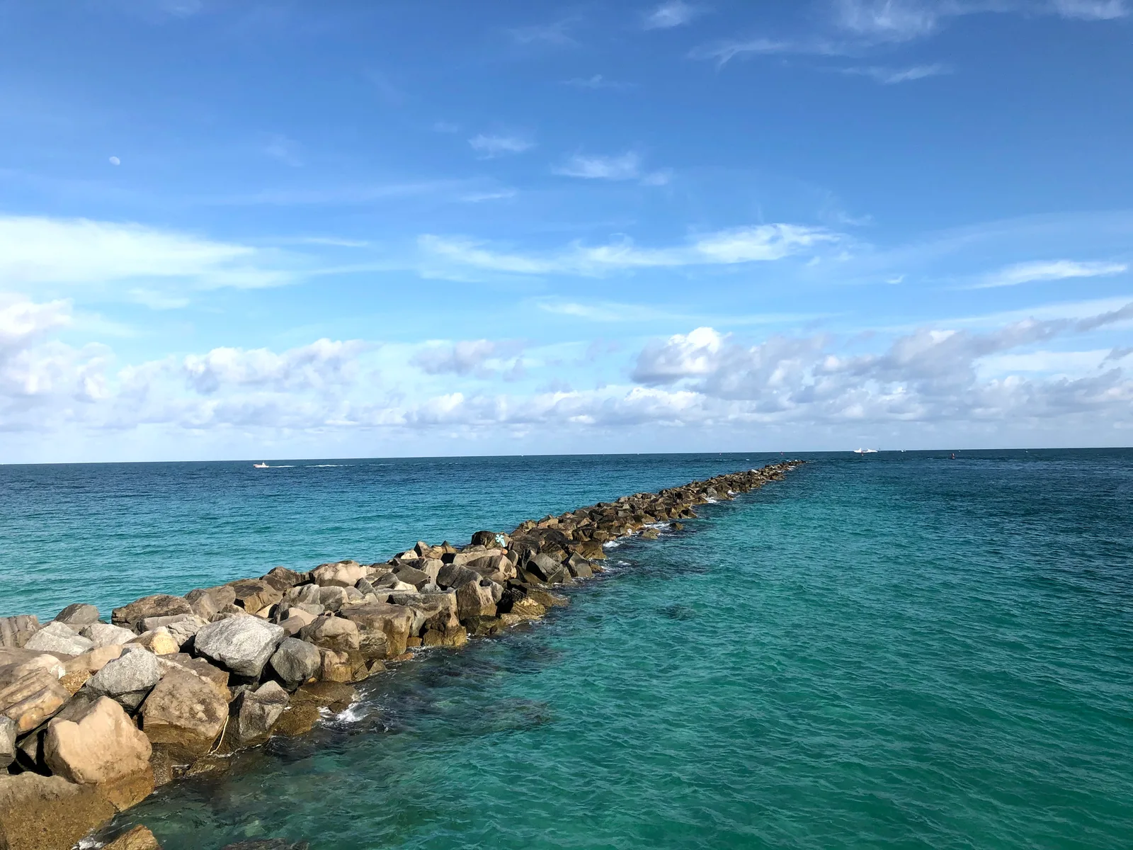 A rock jetty extending into blue ocean water under scattered clouds and a faint moon.