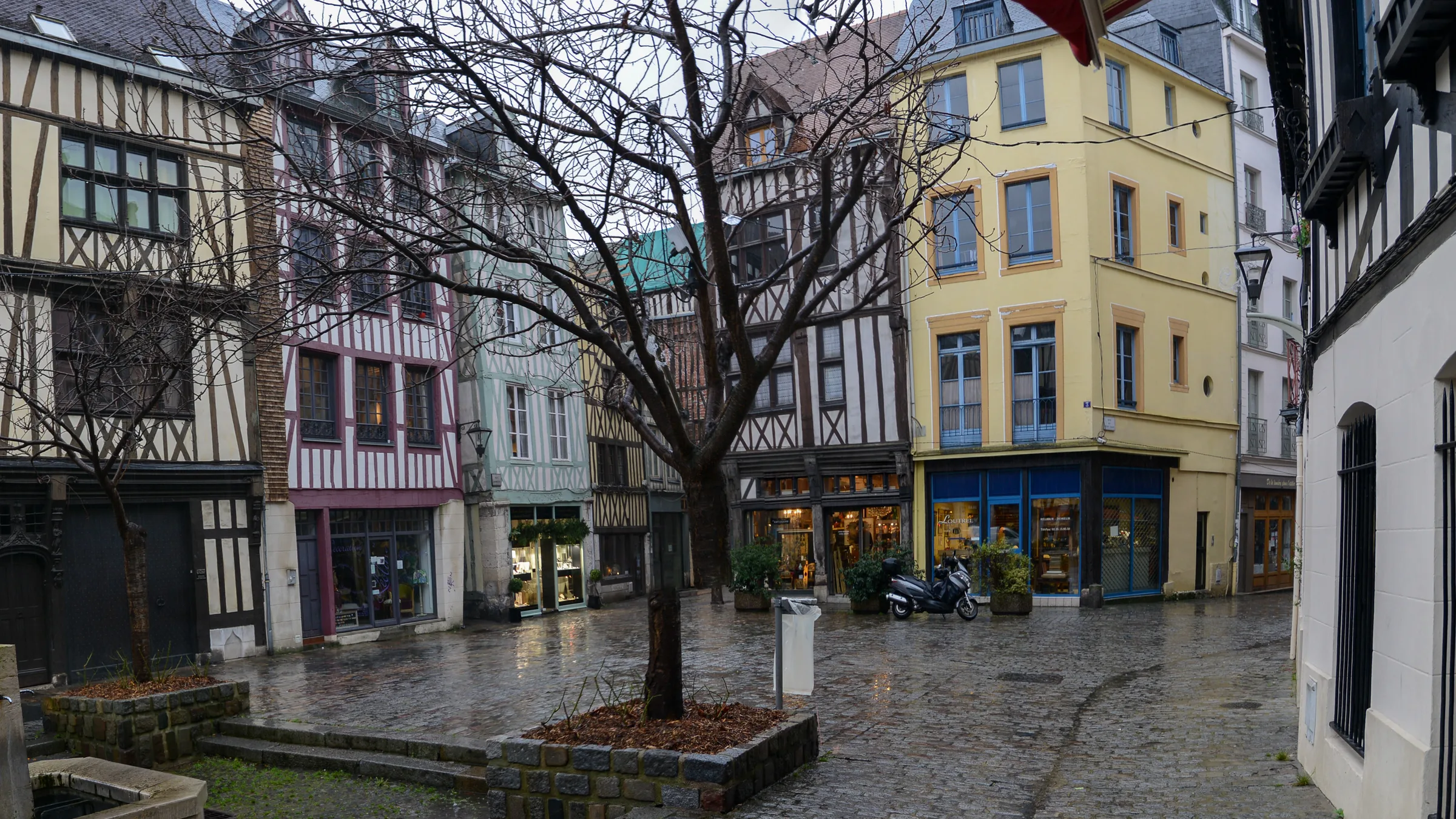 Half-timbered houses around a small paved square with a fountain basin and a bare tree in Rouen's old town after rain.