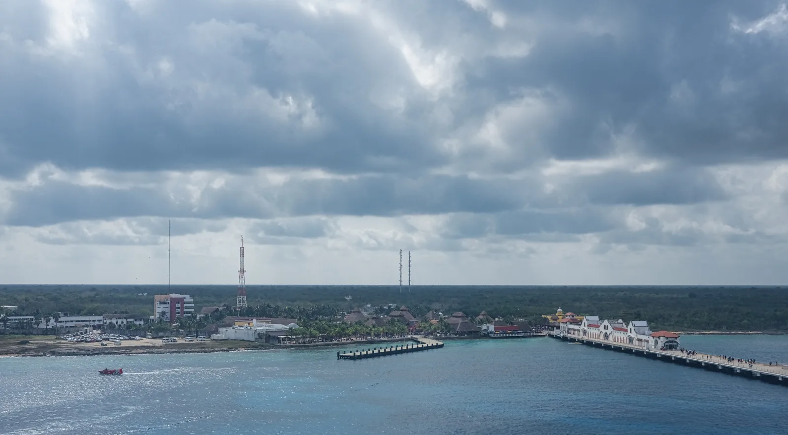 The Cozumel waterfront, port towers and a long cruise pier seen across the water on a cloudy day.