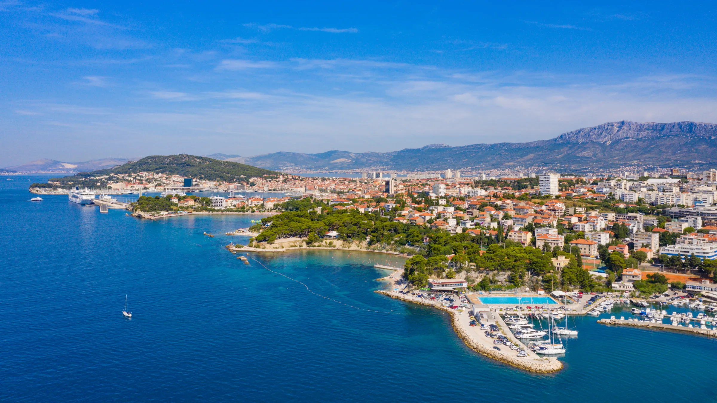 Aerial view over the Bacvice and Firule coast of Split with a swimming pool, a marina and the city rising to the mountains behind.