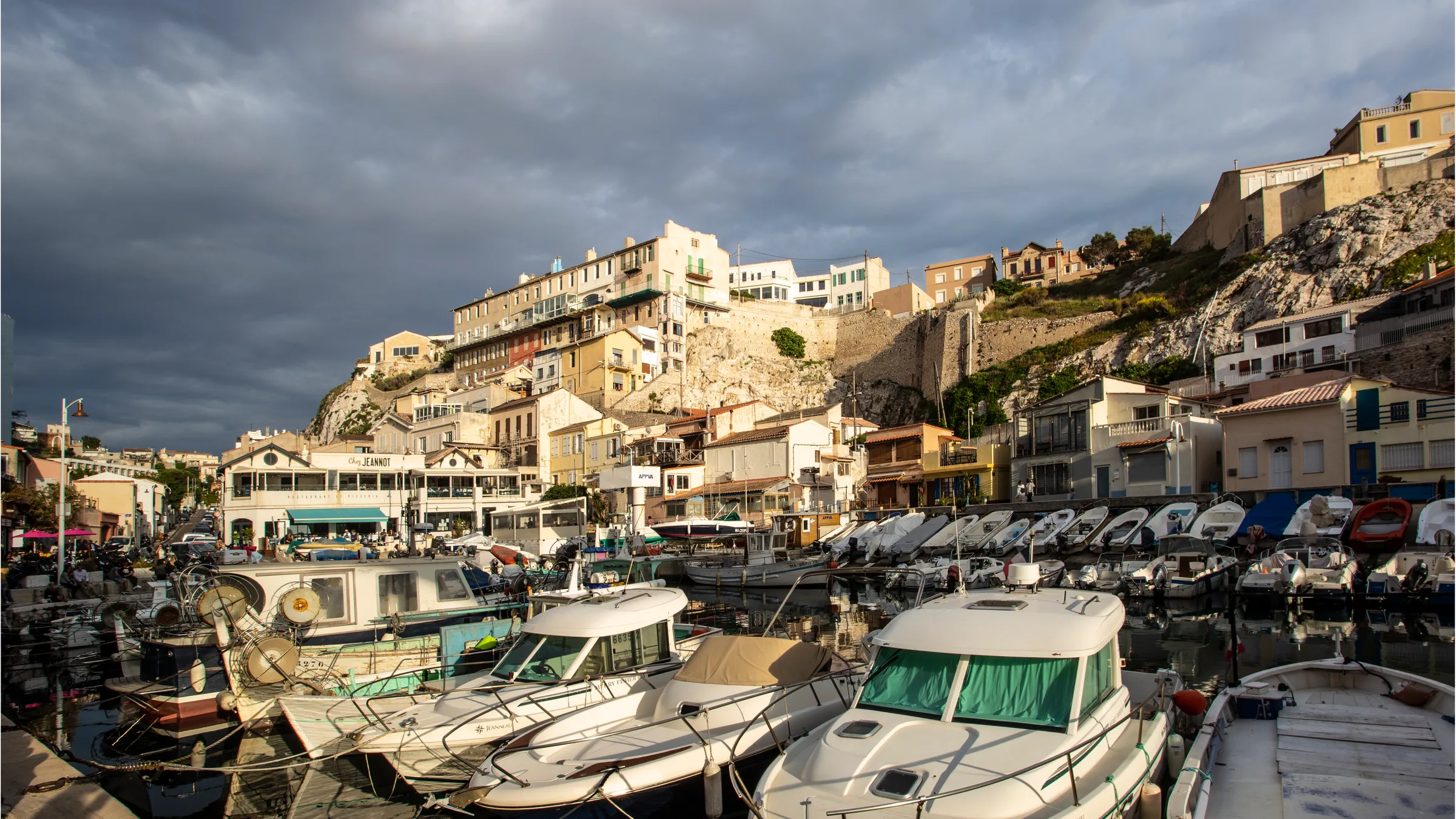 Small fishing and leisure boats packed into the Vallon des Auffes harbour below pastel apartment blocks under a stormy sky.