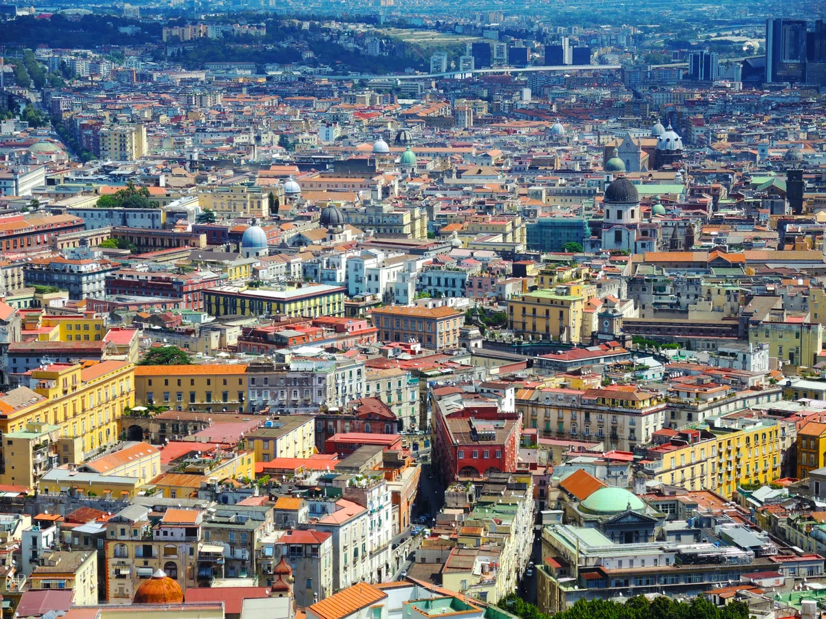A dense, colourful view over central Naples rooftops, domes and the modern business district beyond.
