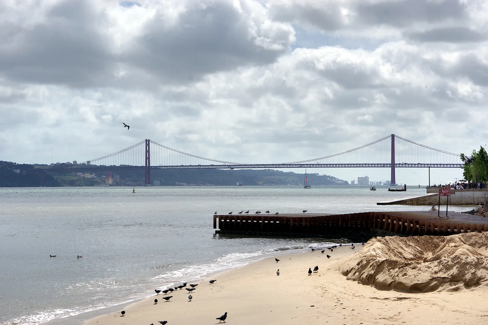 Looking along a sandy riverside beach in Lisbon at the long red 25 de Abril Bridge across the Tagus.