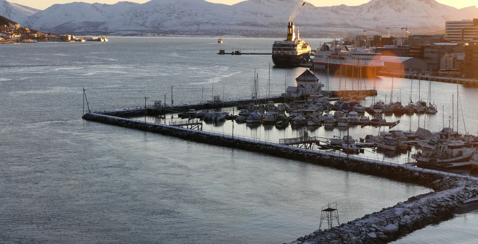 The Tromso waterfront and breakwater at dawn, with a cruise ship at the quay and snowy mountains behind.