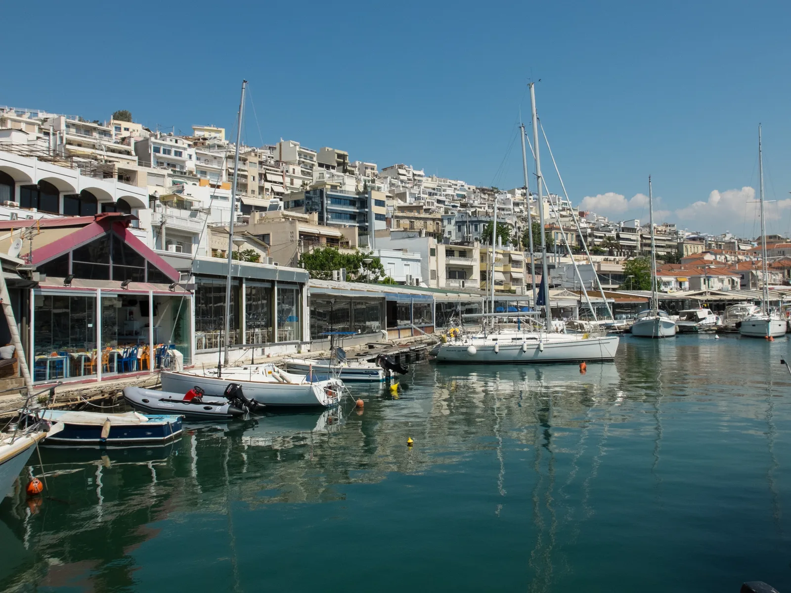 Sailing boats and cafes along the Mikrolimano marina in Piraeus with apartment blocks rising behind.