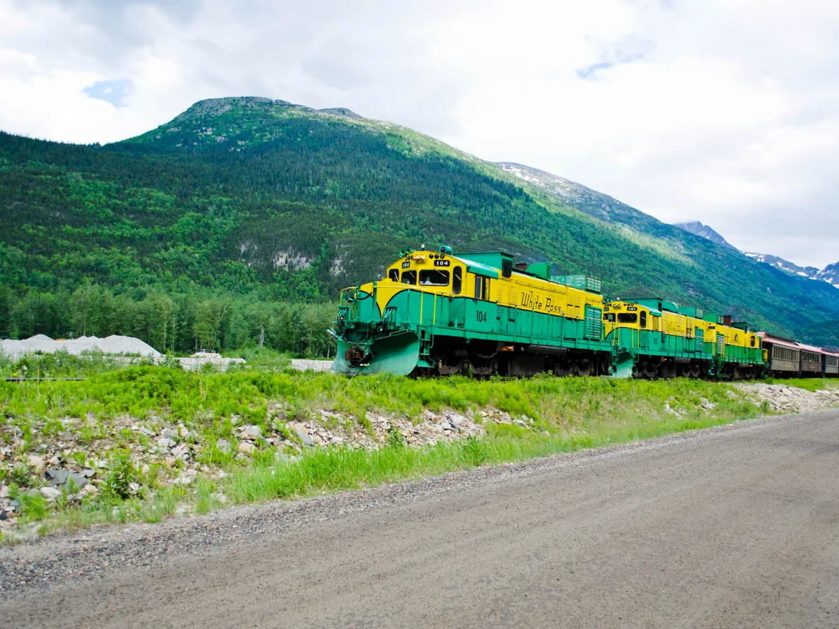 A green and yellow White Pass and Yukon Route diesel train passing along a gravel road below green mountains.